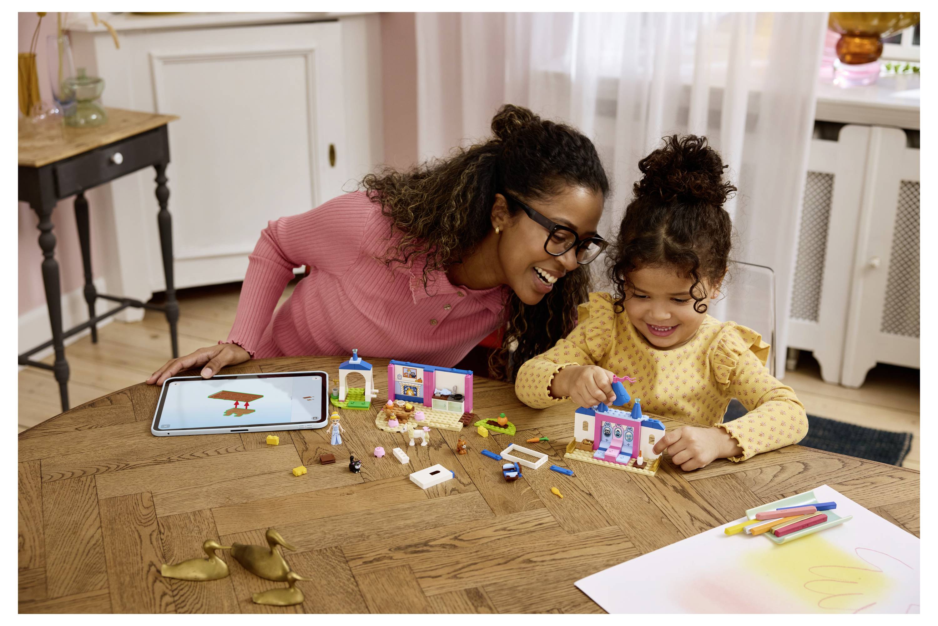 Frau und junges Mädchen lächelnd, während sie an einem Tisch mit Spielzeugblöcken bauen, mit einem Tablet und verstreuten Bastelmaterialien in der Nähe.