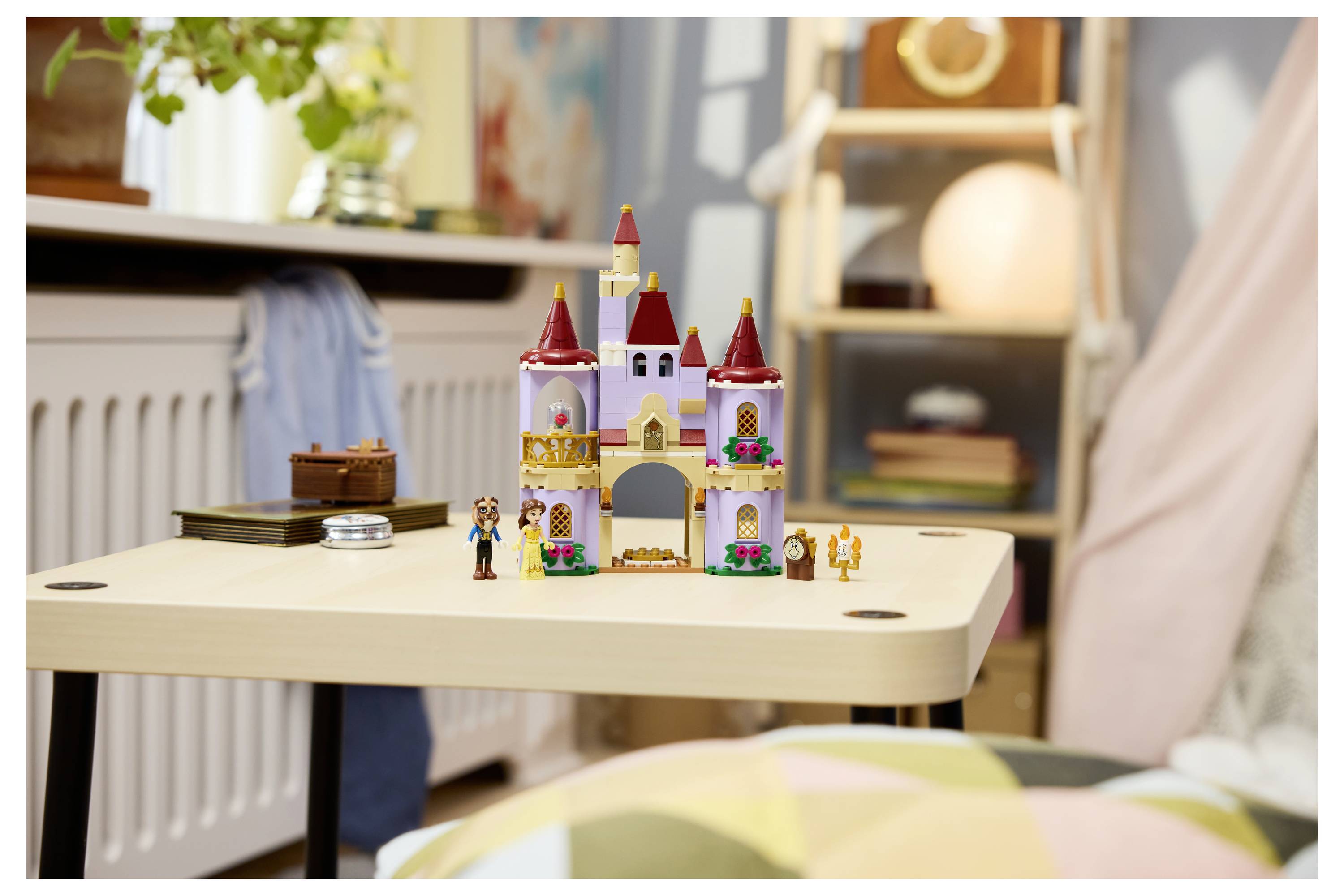 Ein kleines Spielzeugschloss mit Figuren sitzt auf einem Tisch in einem gemütlichen Zimmer, mit einem Stuhl, Büchern und weichen Decken im Hintergrund.