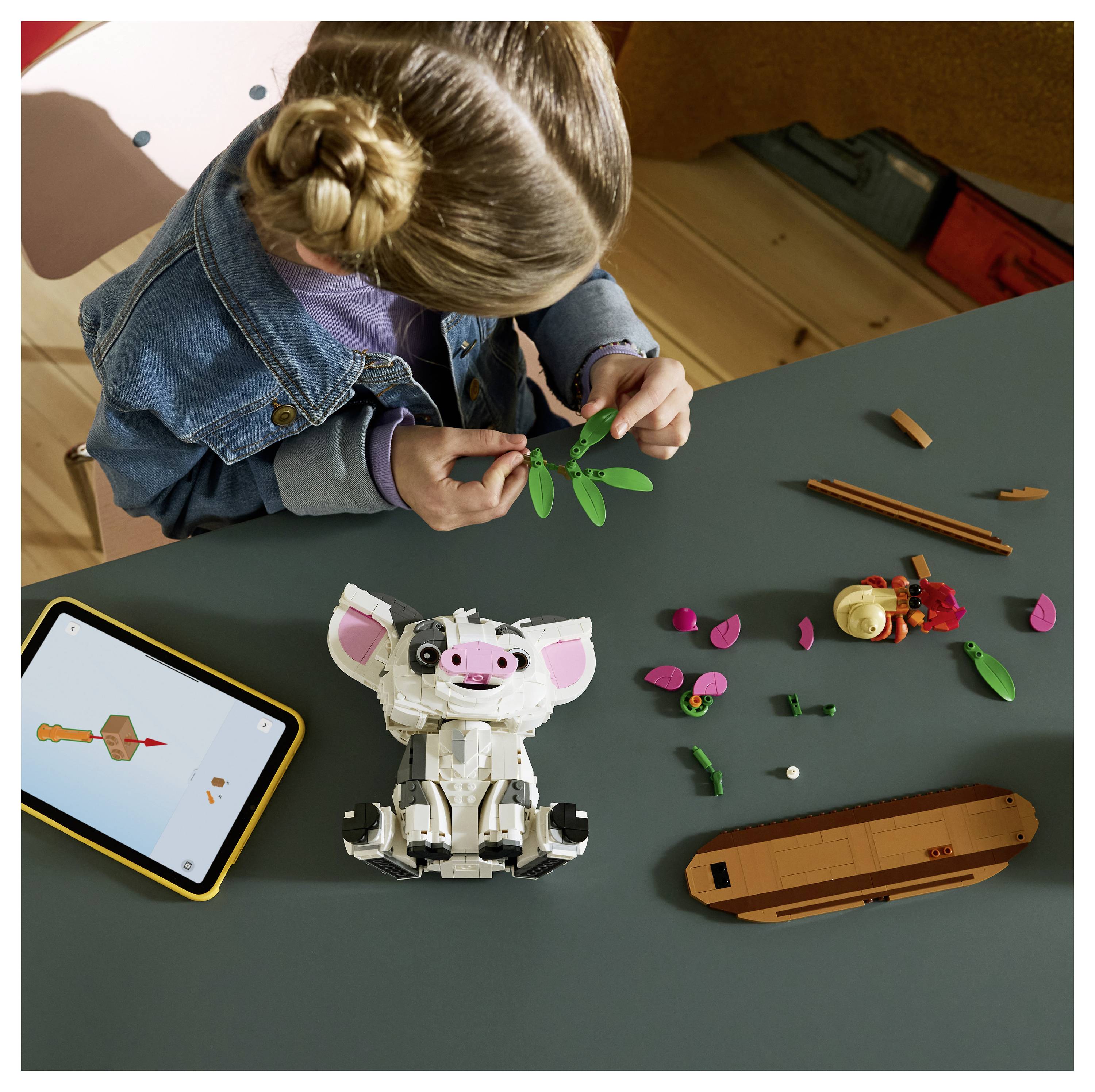 Ein Kind baut ein LEGO-Modell an einem Tisch, mit einem Tablet, einem zusammengebauten Schweinroboter und verstreuten Teilen sowie Werkzeugen in der Nähe.