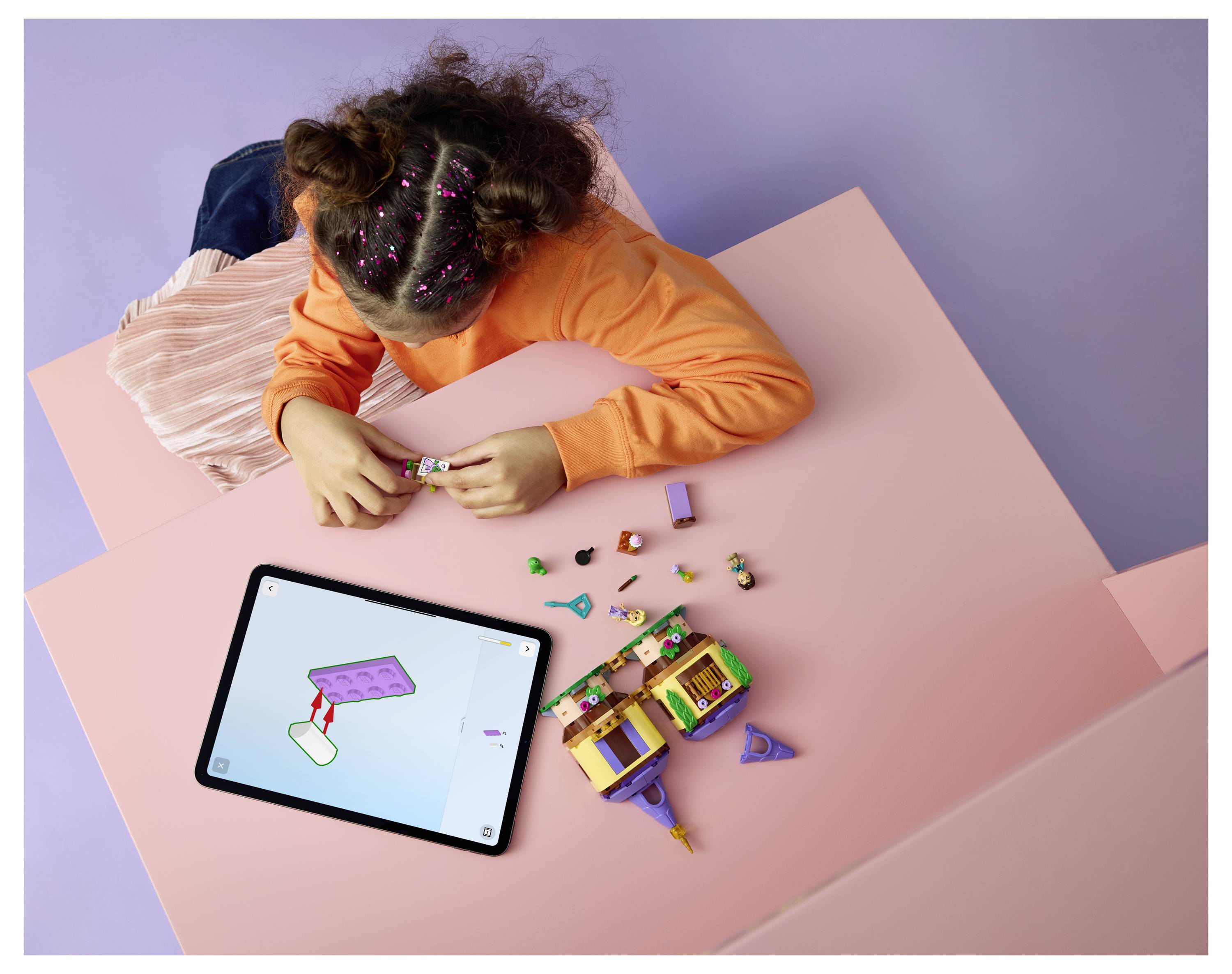 Kind baut ein kleines buntes Spielzeugset auf einem rosa Tisch auf, während es ein Tablet als Anleitung nutzt.