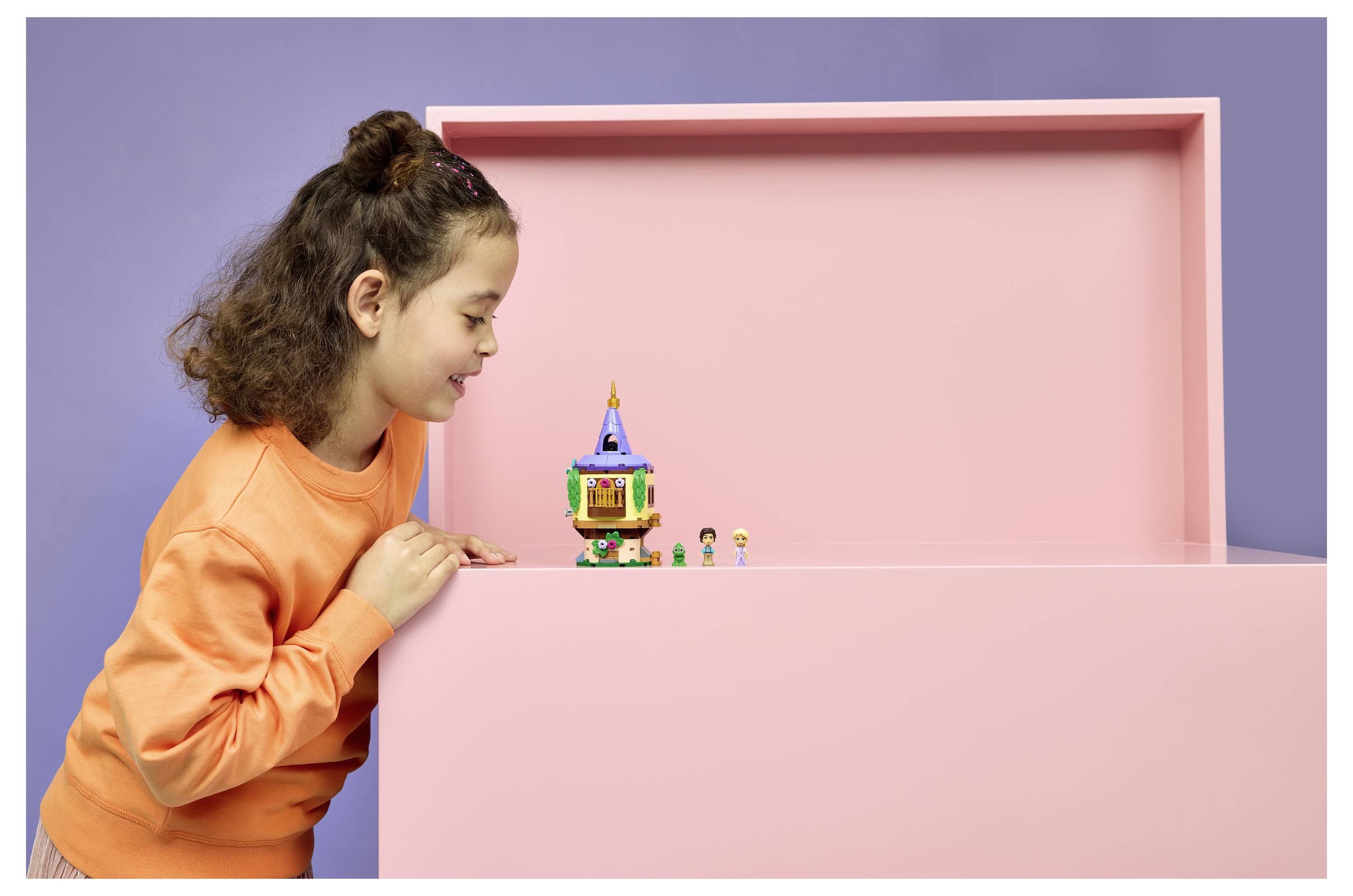 Junges Mädchen in einem orangefarbenen Sweatshirt lehnt sich über eine rosa Schachtel und lächelt auf ein kleines buntes Spielzeuggebäude mit winzigen Figuren.