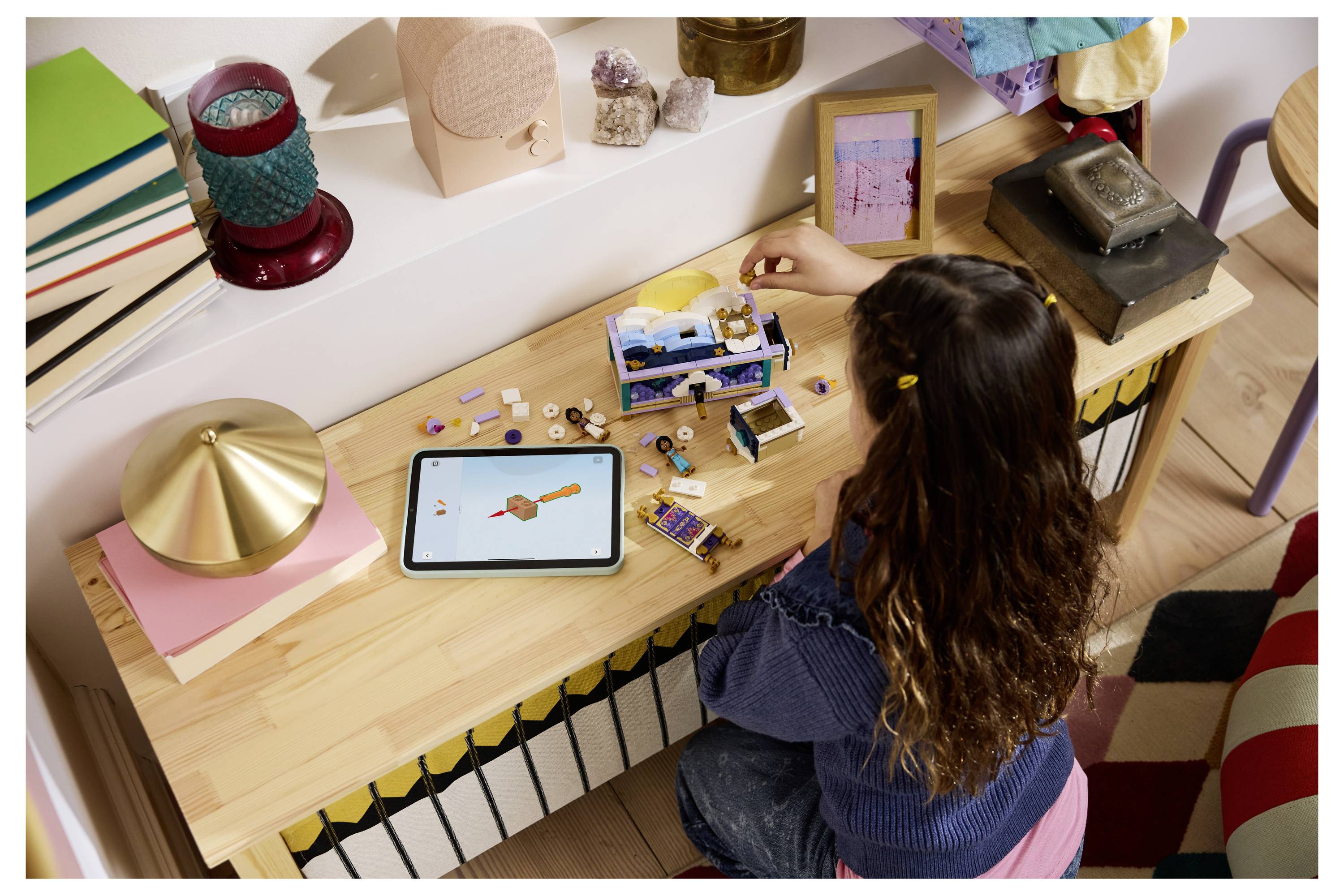 Person, die ein kleines LEGO-ähnliches Modell an einem Schreibtisch neben einem Tablet mit einer Design-Skizze zusammenbaut, mit verstreuten Spielzeugtteilen und Werkzeugen.