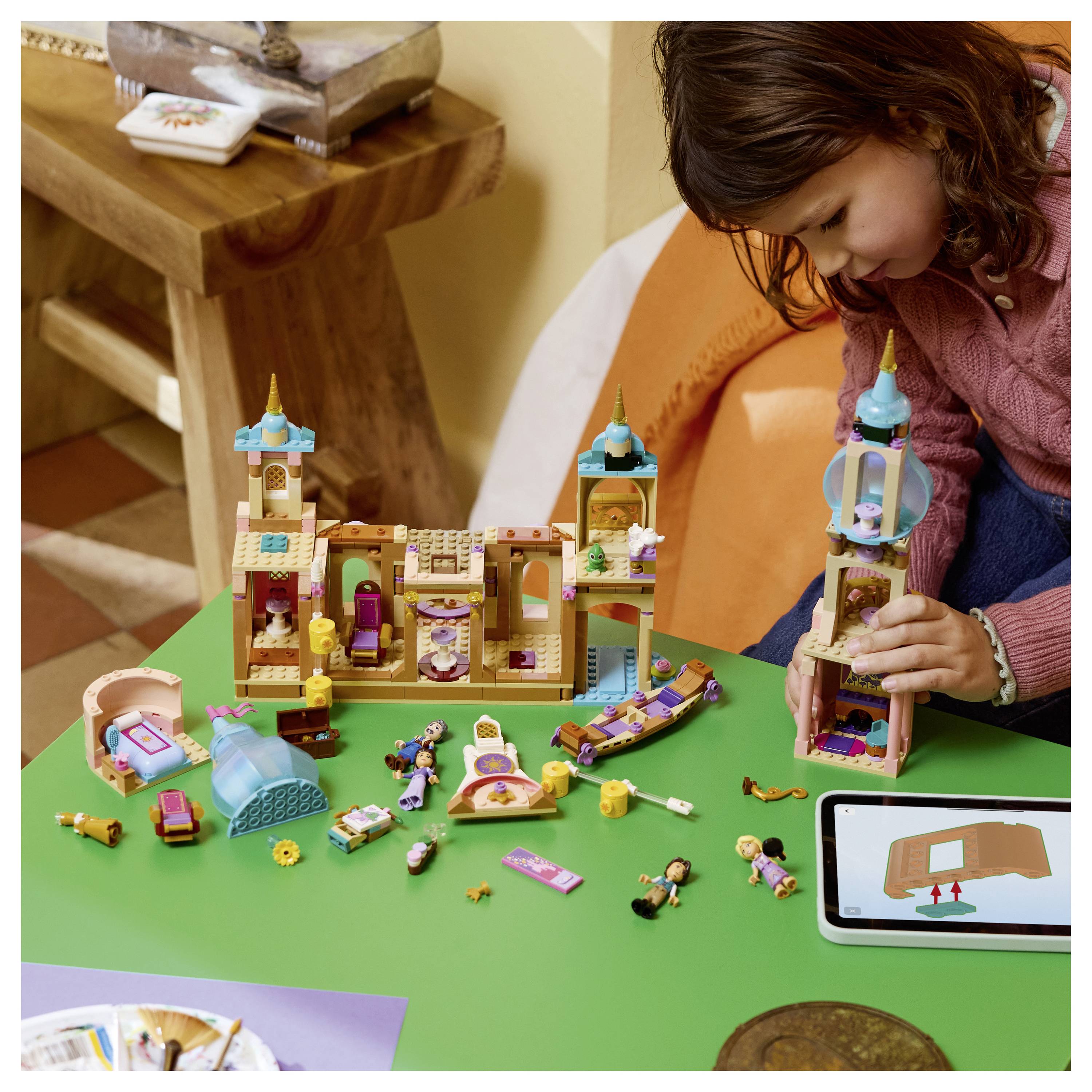 Ein junges Mädchen baut ein farbenfrohes LEGO-ähnliches Schloss auf einem grünen Tisch auf, während ein Tablet die gleiche Anleitung zum Zusammenbau zeigt.