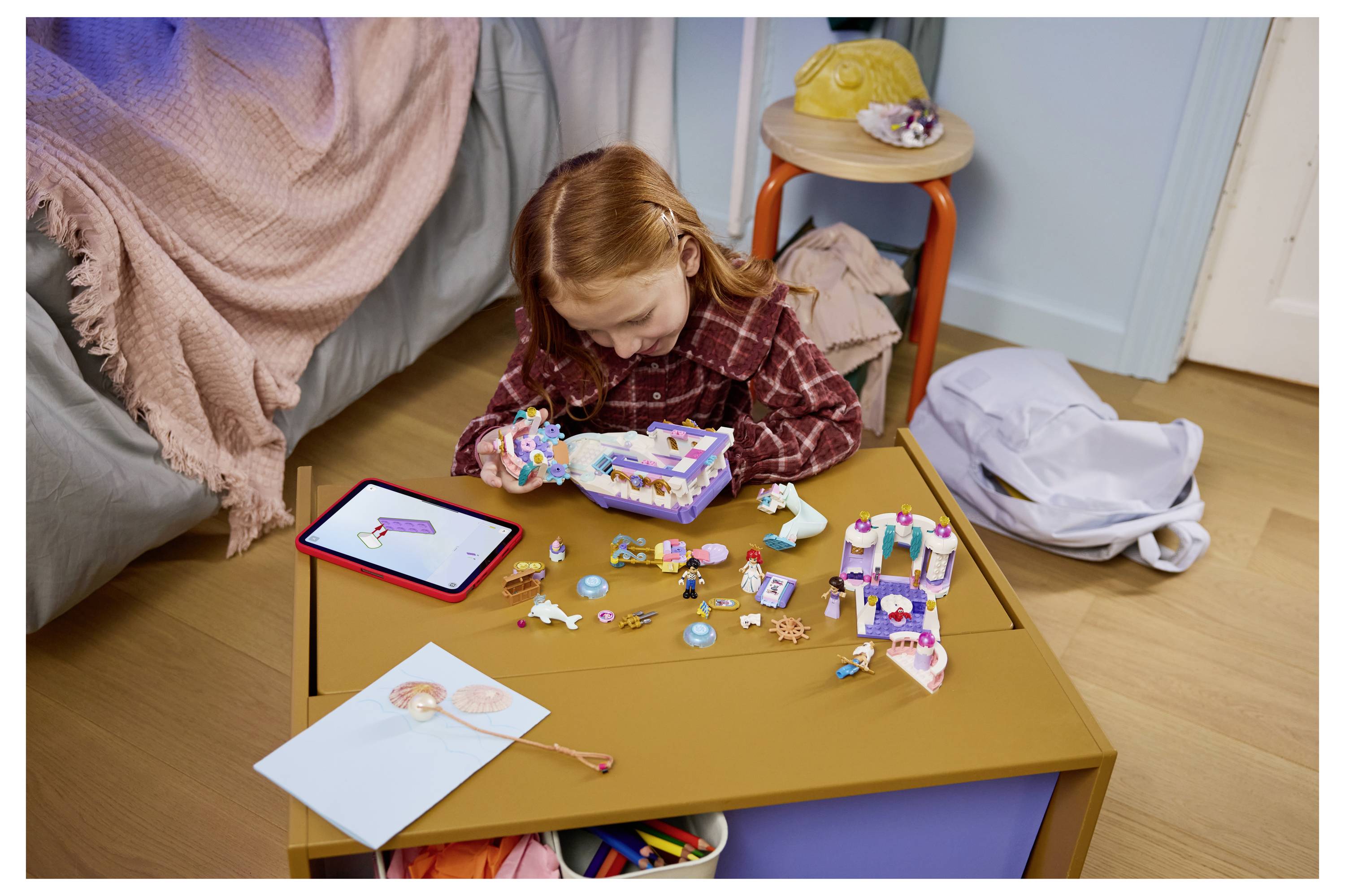 Junges Mädchen sitzt auf dem Boden und montiert ein buntes Spielzeugset, umgeben von verstreuten Teilen, einem Tablet und Bastelmaterialien auf einem kleinen Tisch.