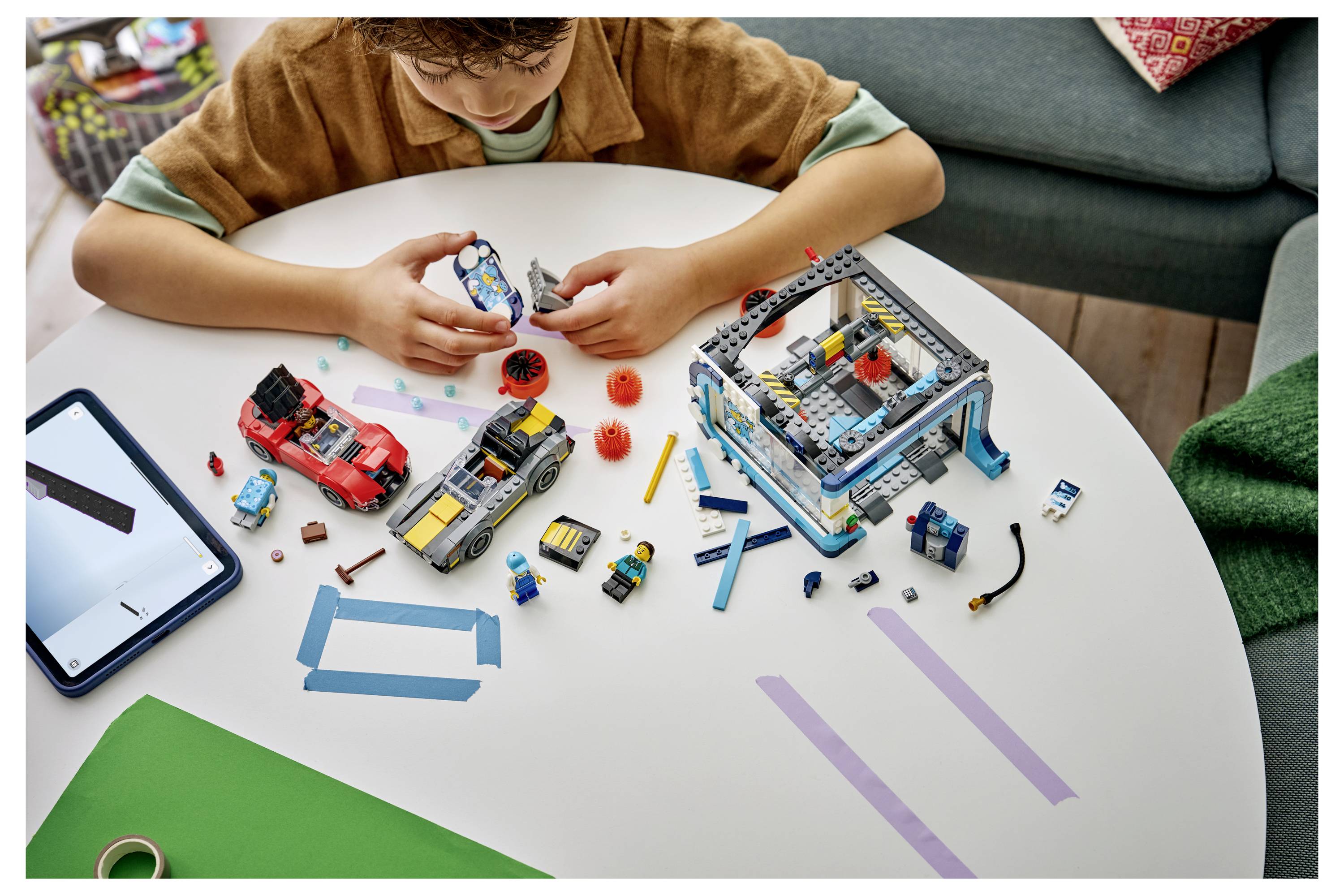 Kind, das ein LEGO-ähnliches Modell an einem Tisch zusammenbaut, mit verstreuten Bausteinen, Anleitung auf einem Tablet und teilweise zusammengebauten Spielzeugfahrzeugen und Strukturen.