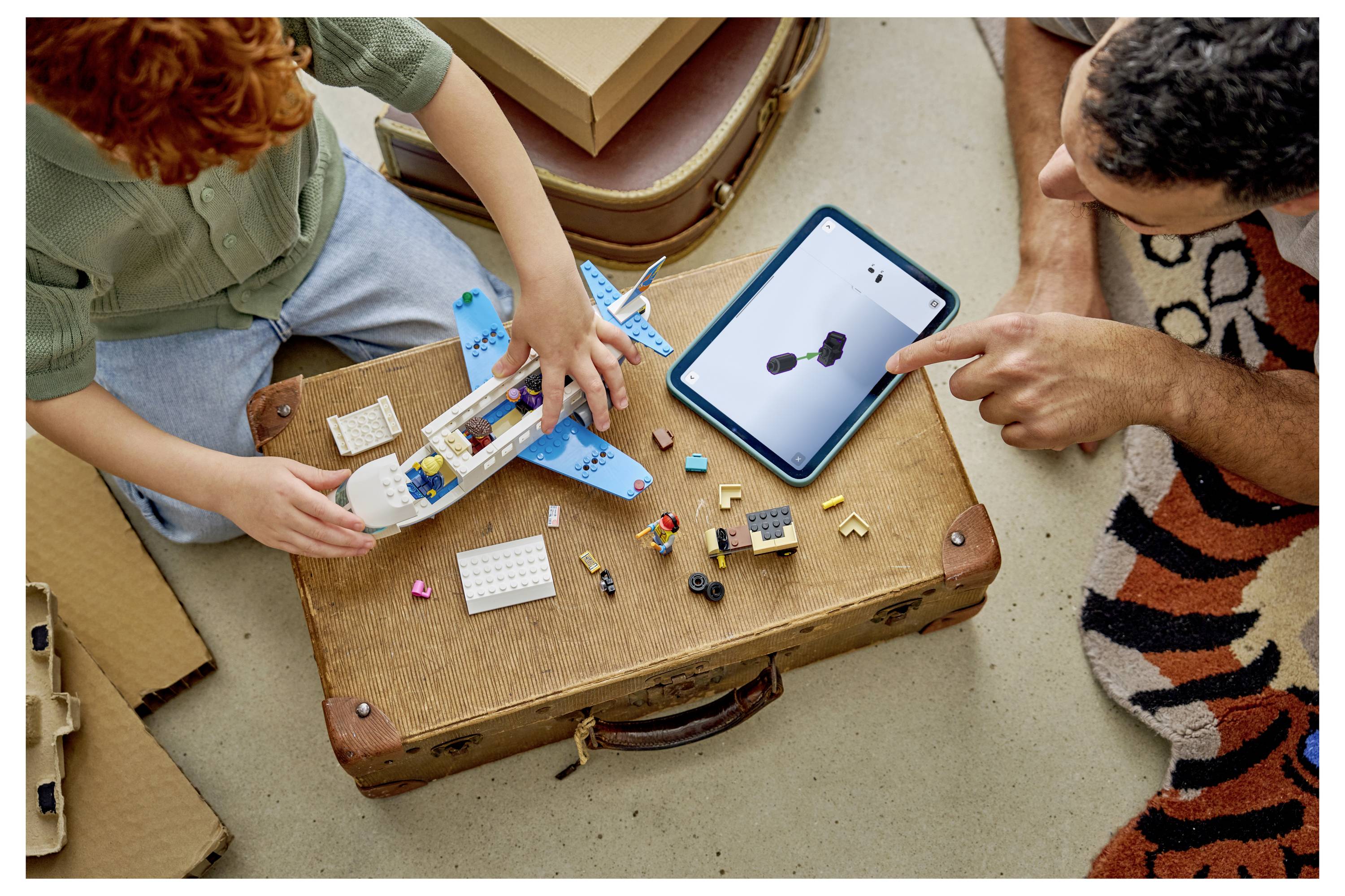 Zwei Personen und ein Kind bauen mit LEGO-ähnlichen Bausteinen ein kleines Spielzeugflugzeug, während eine andere Person auf ein Tablet zeigt, das ein 3D-Modell anzeigt.
