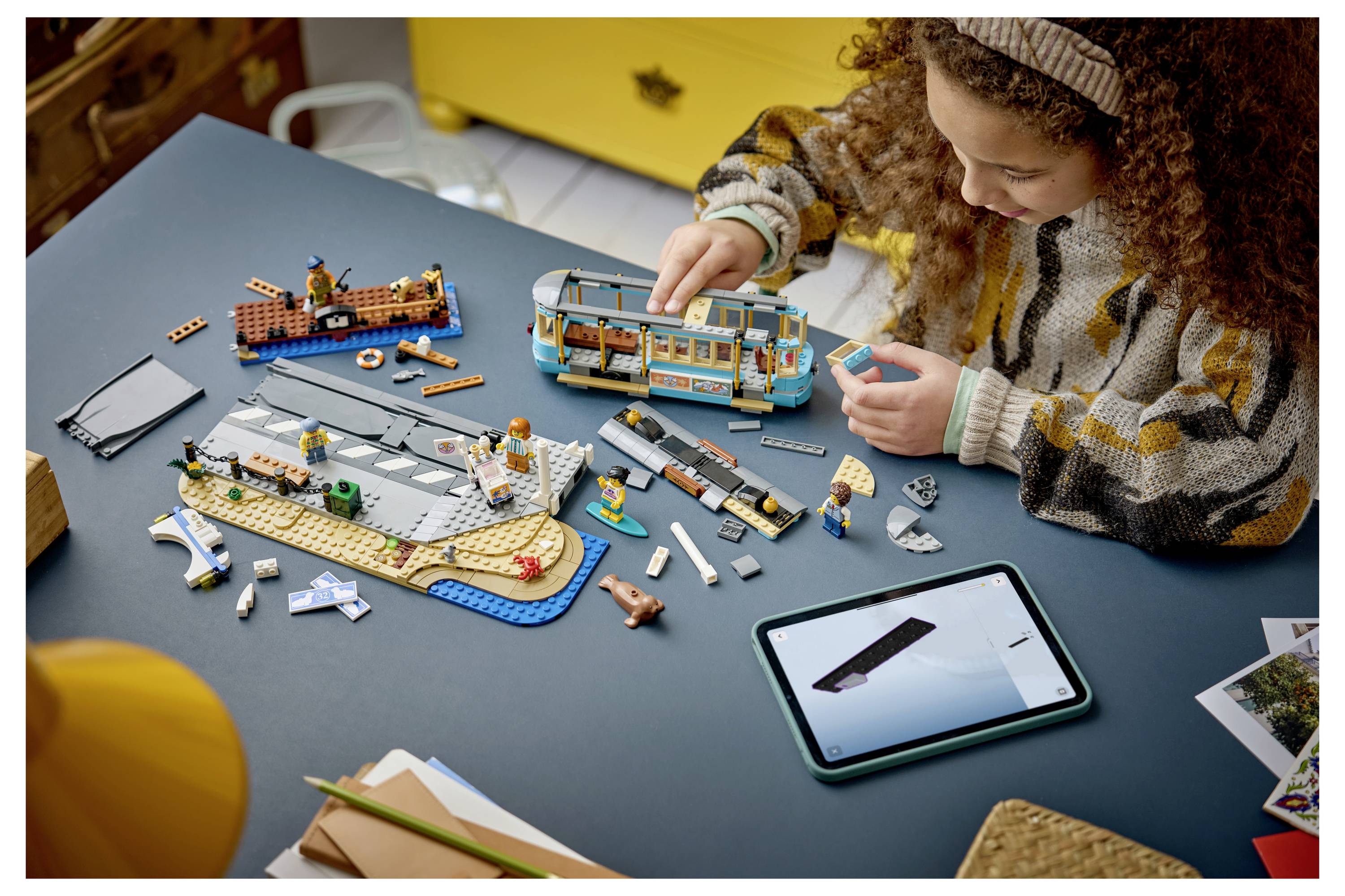 Ein Kind baut ein LEGO-ähnliches Modell auf einem Tisch mit Anweisungen auf einem Tablet, umgeben von verstreuten Teilen und anderen zusammengesetzten Spielzeugteilen.