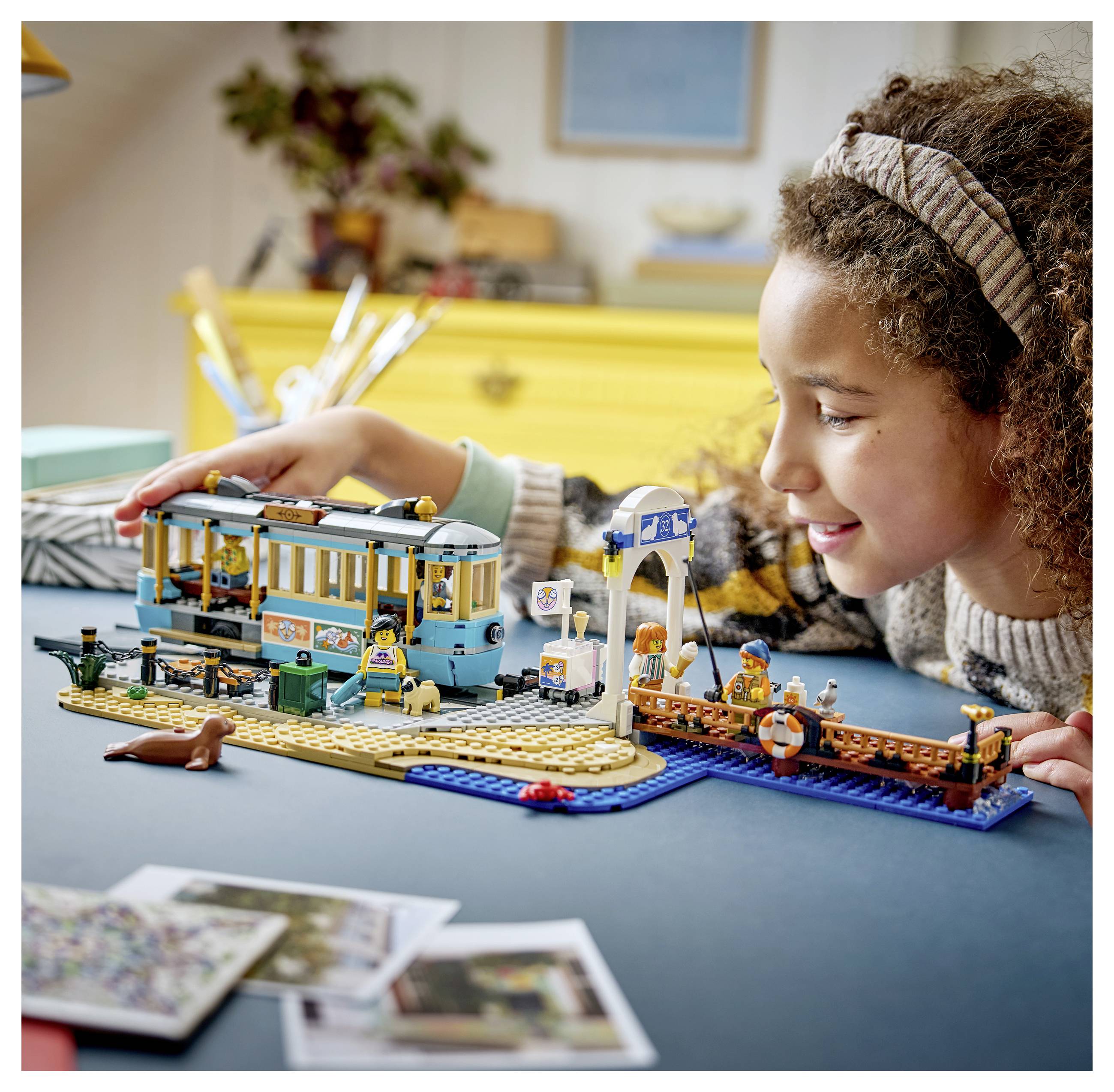 Kind lächelt, während es auf einem Tisch mit einem LEGO-ähnlichen Straßenbahn-Set am Hafen spielt, mit Dock, Pier und Minifiguren.
