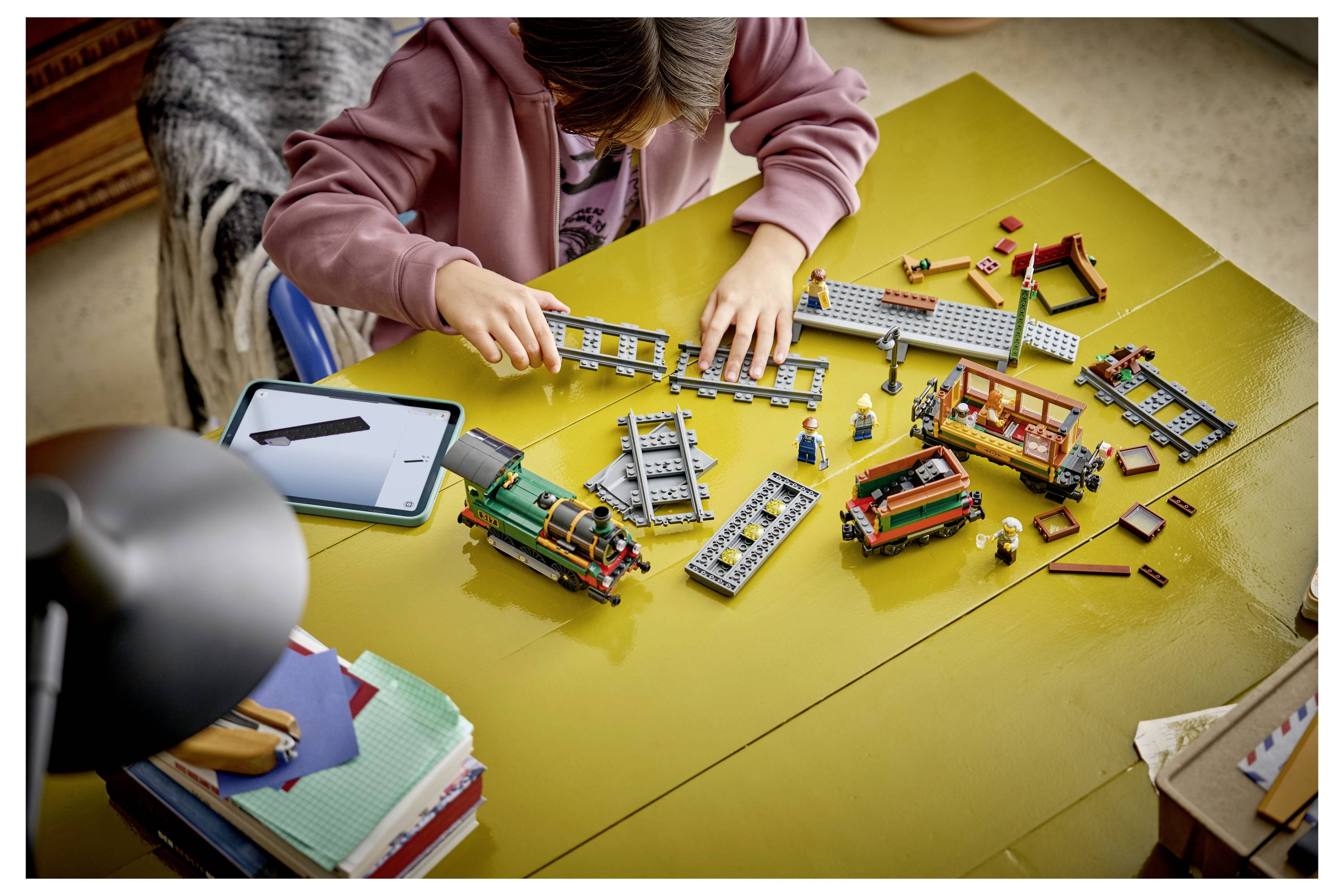 Ein Kind baut ein LEGO-Zugsset an einem gelben Tisch zusammen, mit Gleisen, Zugteilen und kleinen Figuren, die rundherum verteilt sind.