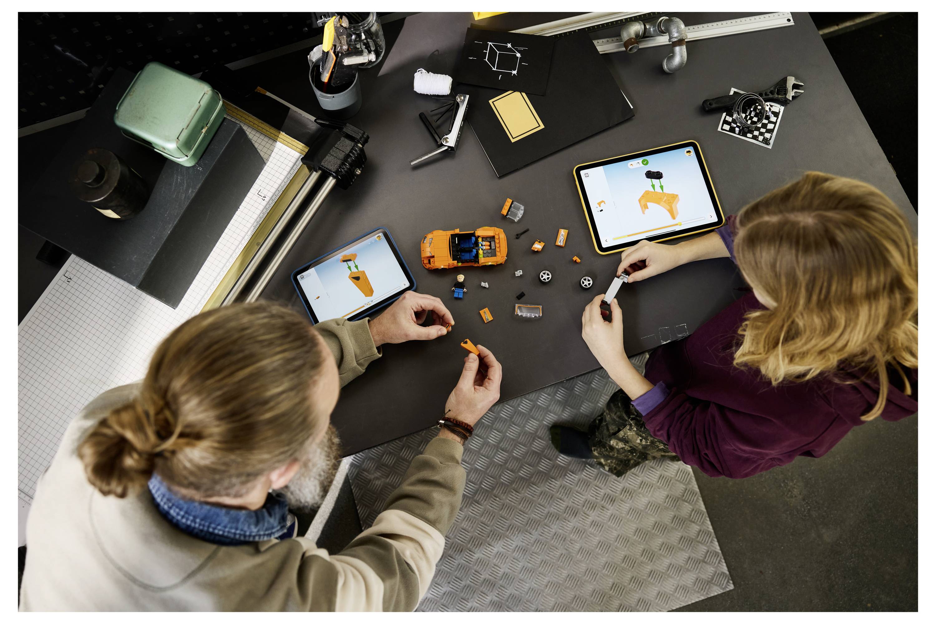 Zwei Personen montieren ein kleines orangefarbenes Spielzeugauto auf einer Werkbank, wobei sie Tablet-Anleitung und verstreute Teile in einer Werkstatt nutzen.
