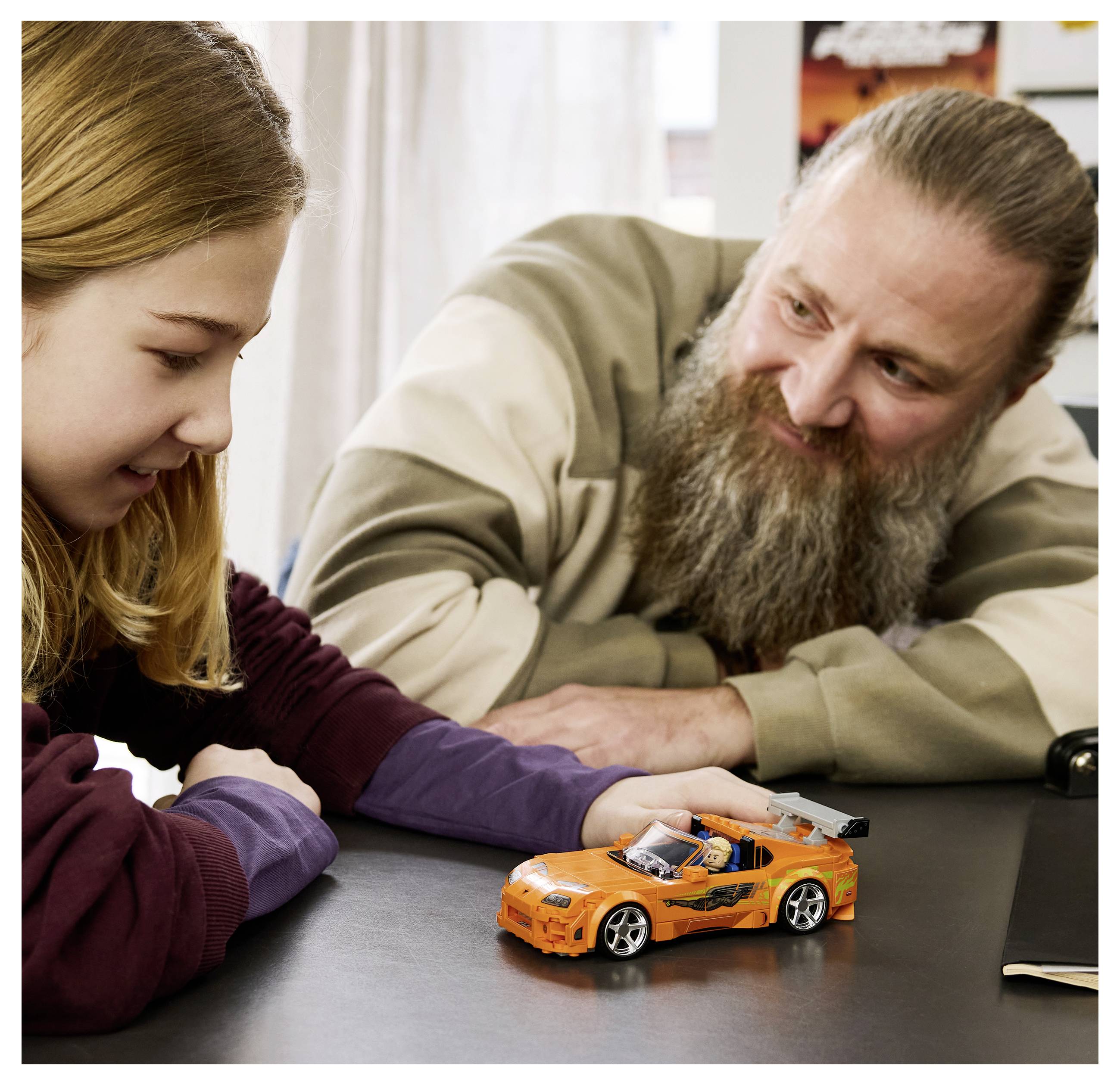 Ein Mann und ein Mädchen lächeln ein orangefarbenes Spielzeugauto auf einem Tisch an, was auf eine verspielte Interaktion oder die Besprechung eines Kinderspielzeugs hindeutet.