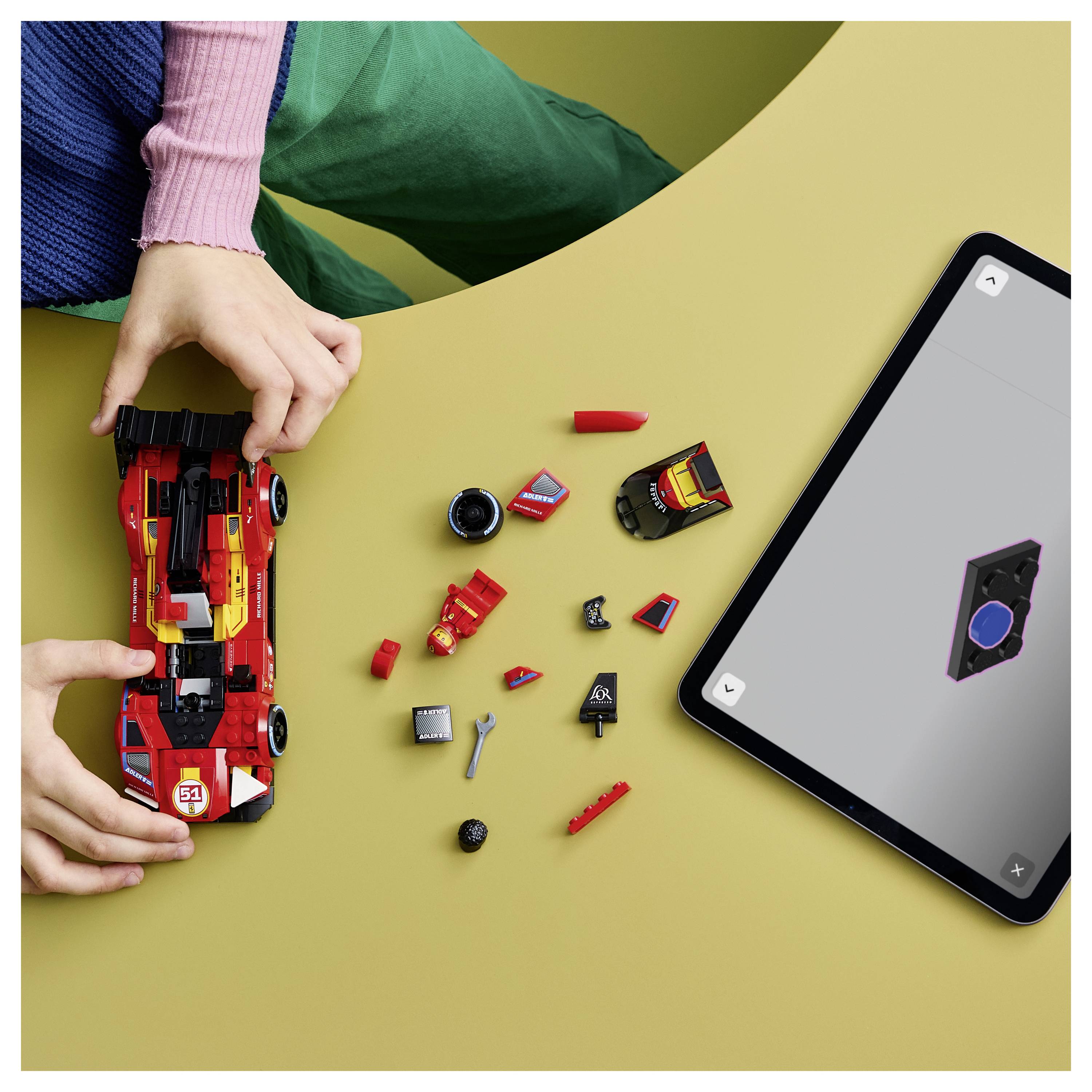 Ein Kind montiert ein rotes Spielzeug-Rennwagen aus LEGO-ähnlichen Bausteinen neben einem Tablet, das das Autodesign auf einer gelben Oberfläche anzeigt.