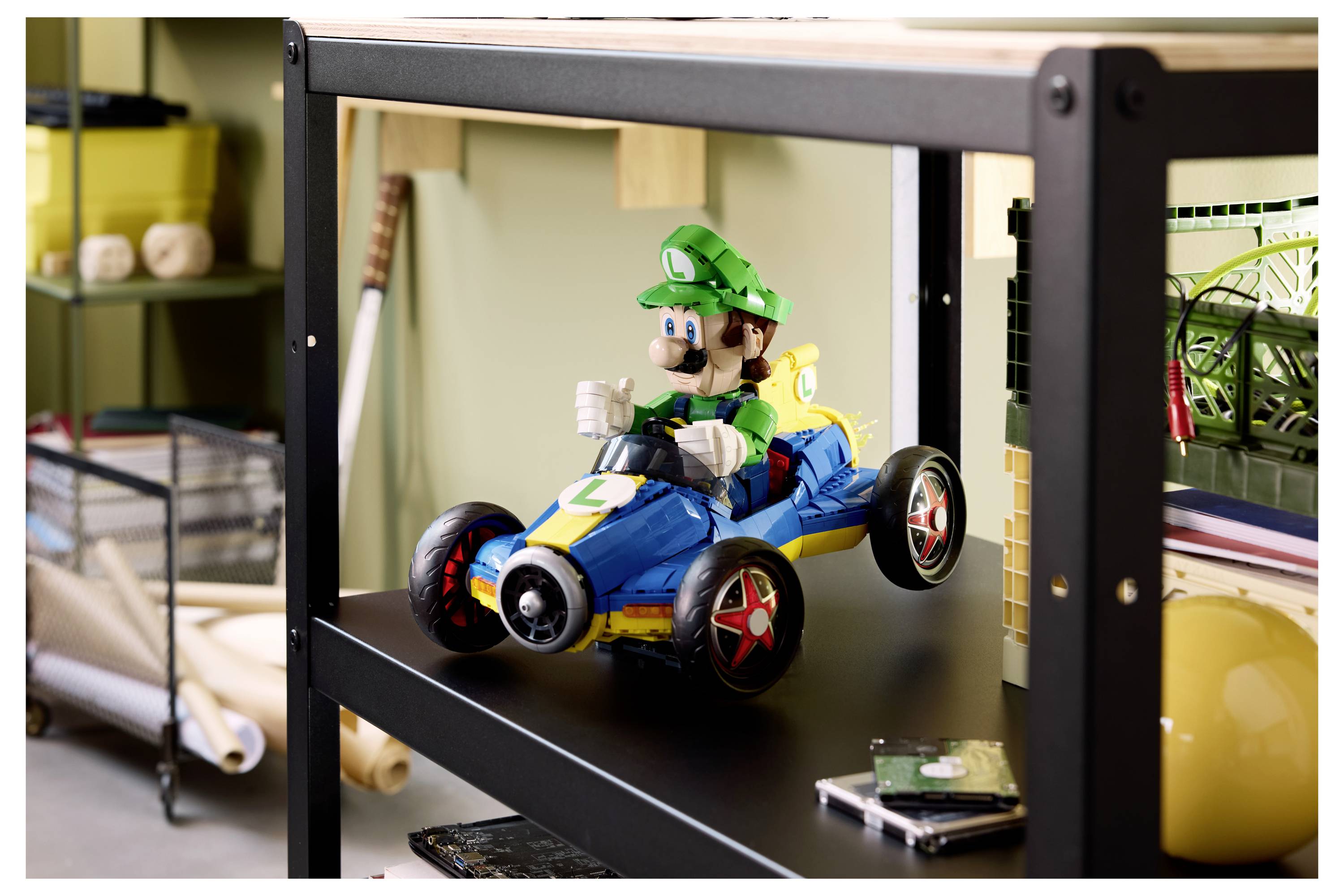 Eine Luigi-Figur fährt in einem blauen Spielzeugkart auf einem schwarzen Regal, mit Bastelmaterialien und Werkzeugen, die um sie herum sichtbar sind.
