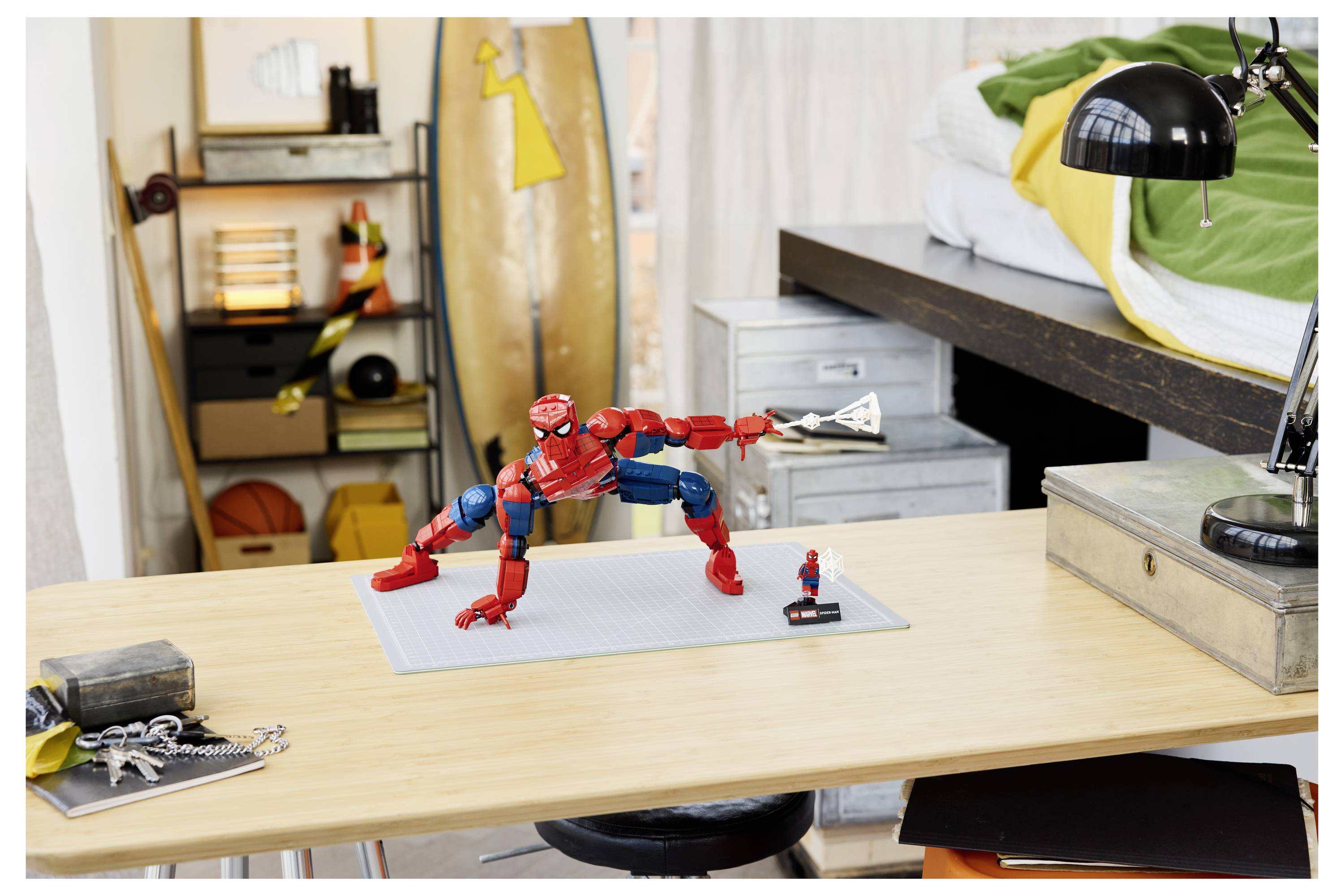 Spider-Man-Actionfigur, die auf einem Schreibtisch in einem Schlafzimmer posiert, mit einer kleinen Minifigur daneben und einem Skateboard sowie einer Lampe im Hintergrund.