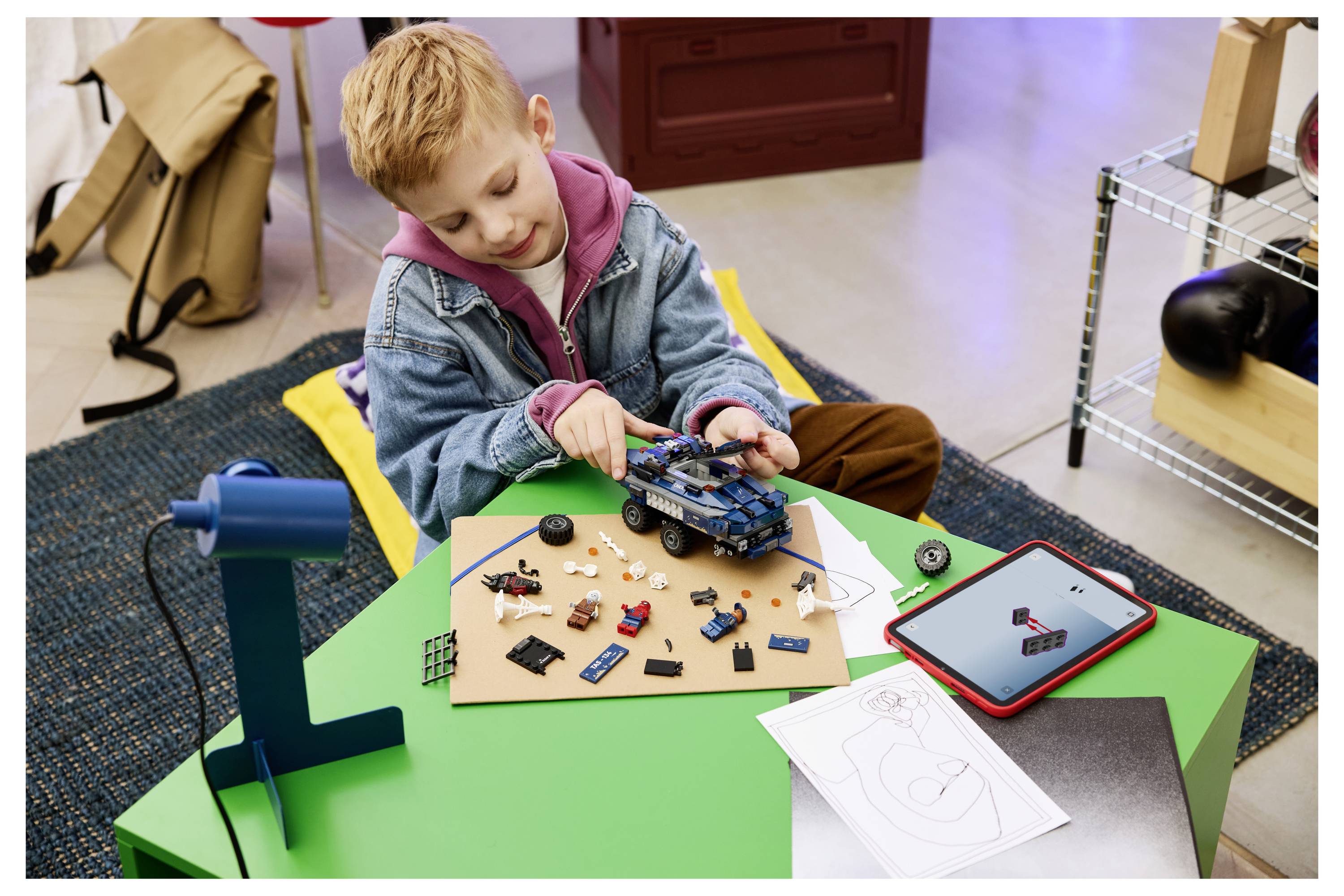 Ein junger Junge montiert ein Spielzeugauto mit elektronischen Teilen an einem niedrigen grünen Tisch, mit einem Tablet und einer Anleitung neben ihm.