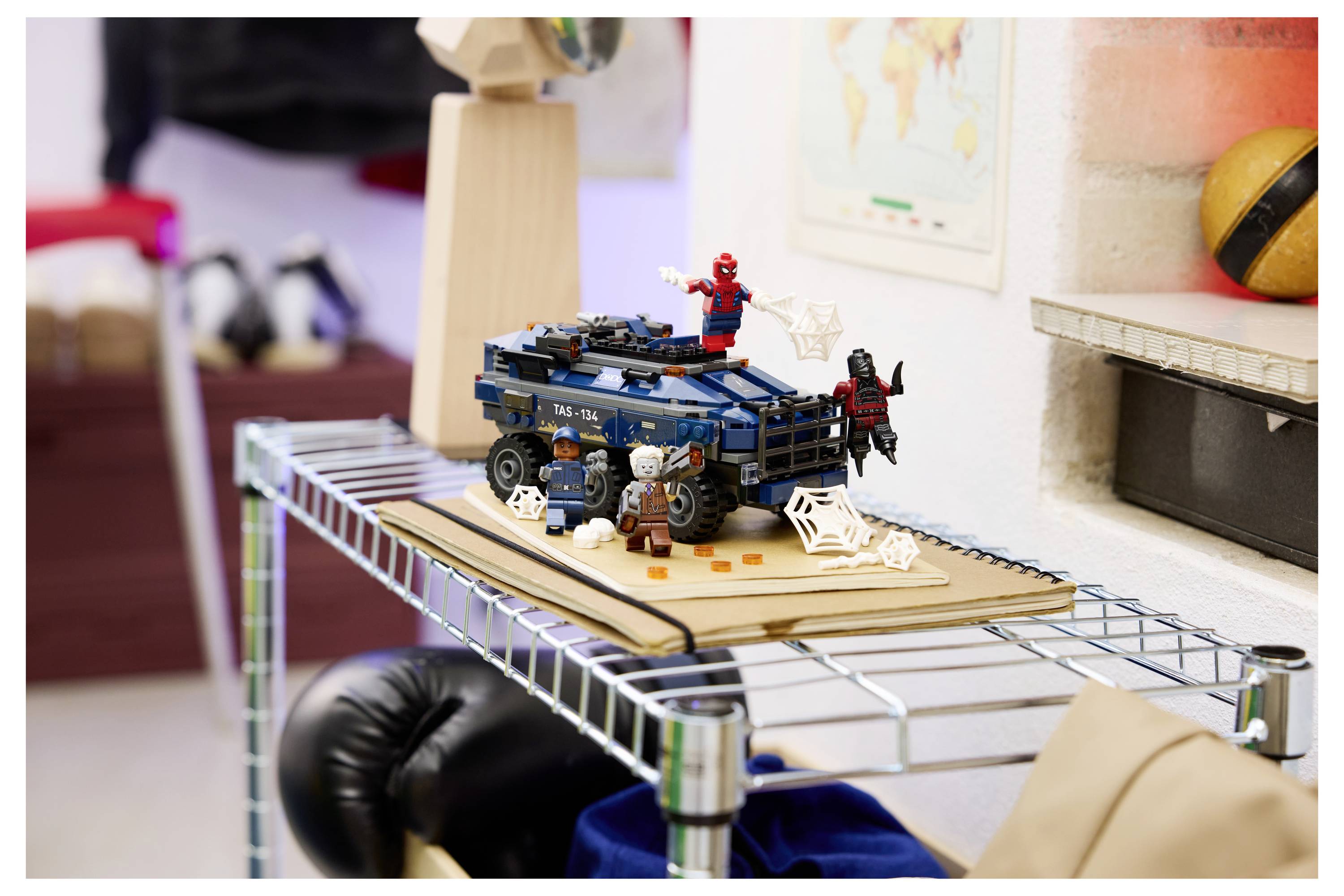 LEGO-Superhelden-Fahrzeugszene auf einem Regal, mit Spider-Man und einer anderen Figur, die auf einem blauen Auto posiert werden, umgeben von Spinnennetz-Dekorationen.