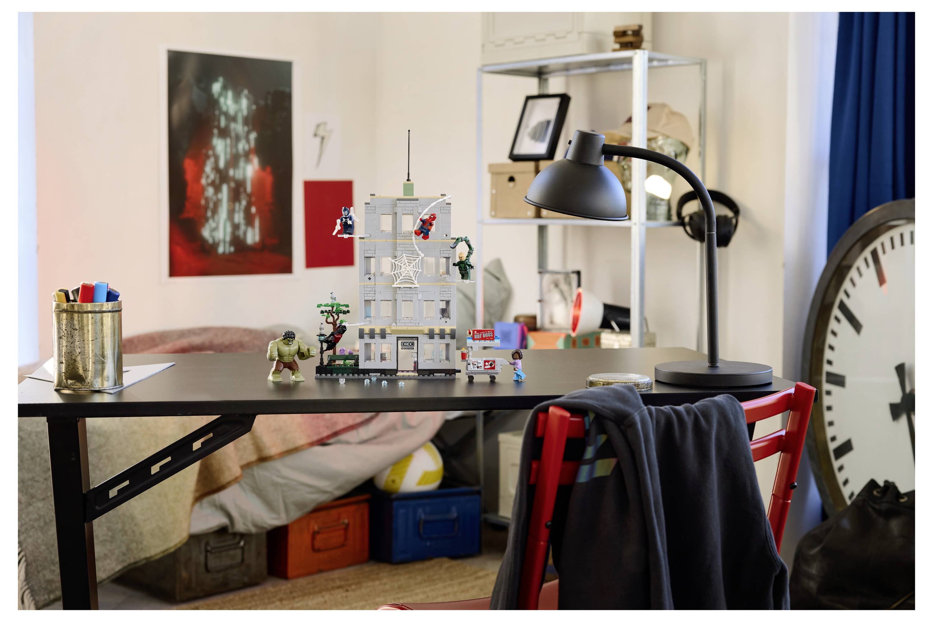 Ein Kinderzimmer mit einem Schreibtisch, der mit LEGO-Steinen und einer gebauten Struktur bedeckt ist, einer Lampe, Regalen und einer großen Uhr am Fenster.