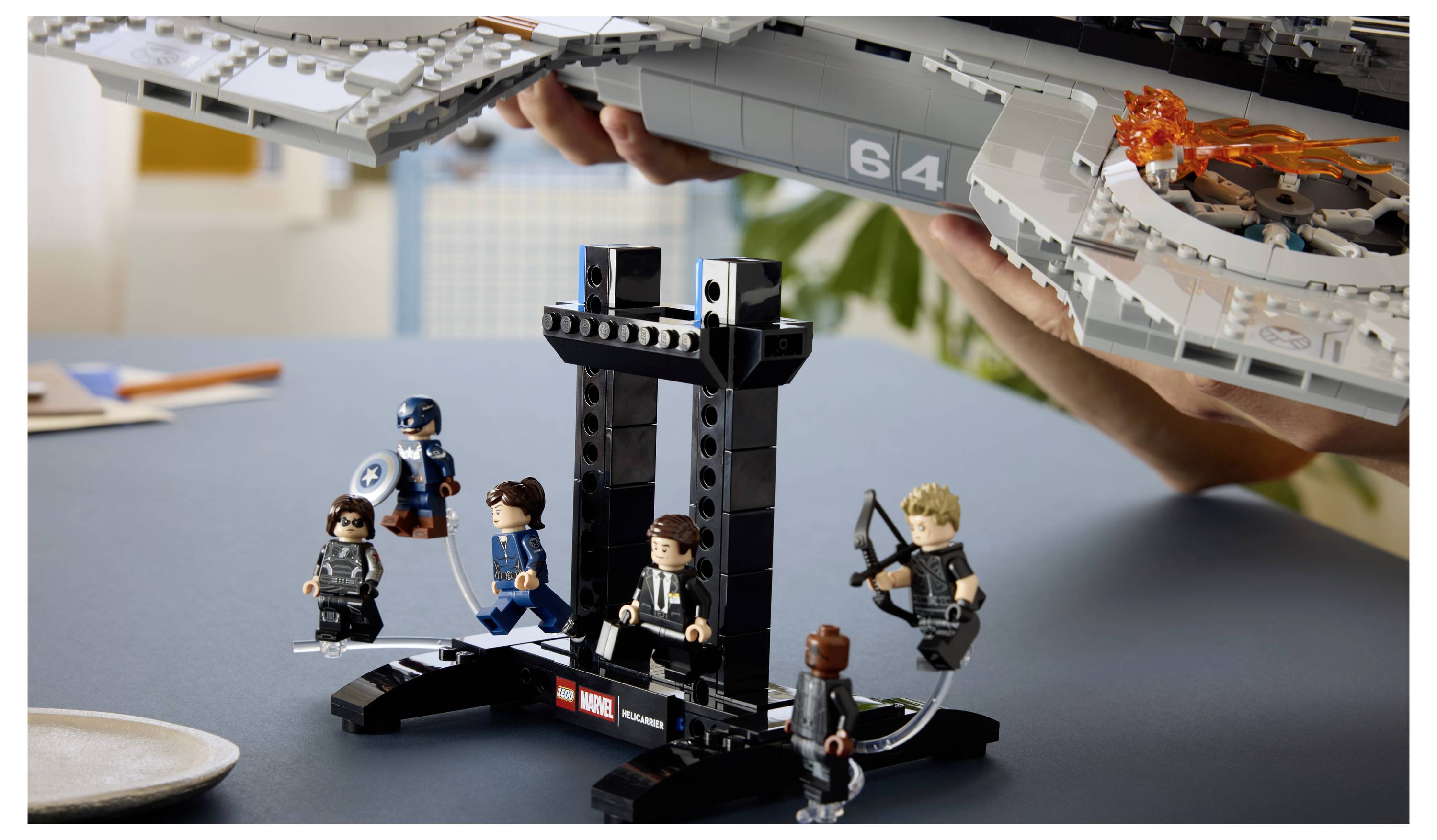 LEGO Marvel Ausstellung mit fünf Minifiguren um einen schwarzen Ständer herum, während Hände ein großes graues Schiff über dem Kopf halten, mit einem kleinen orangefarbenen Explosionsdetail auf dem Schiff.