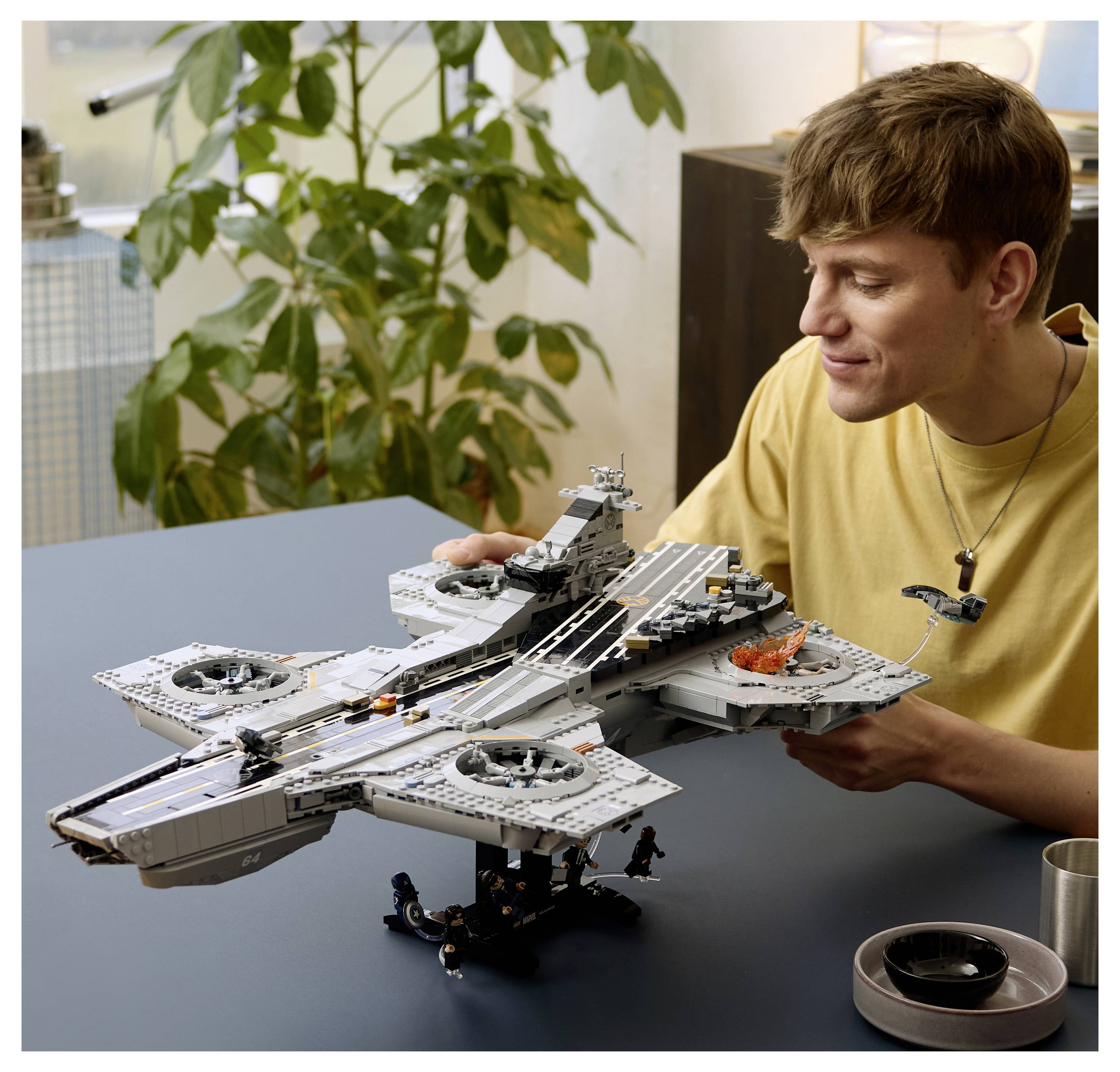 Junger Mann in einem gelben Hemd, der ein großes graues LEGO-ähnliches Flugzeug- oder Raumschiffmodell auf einem Tisch in einem Innenraum zusammenbaut.