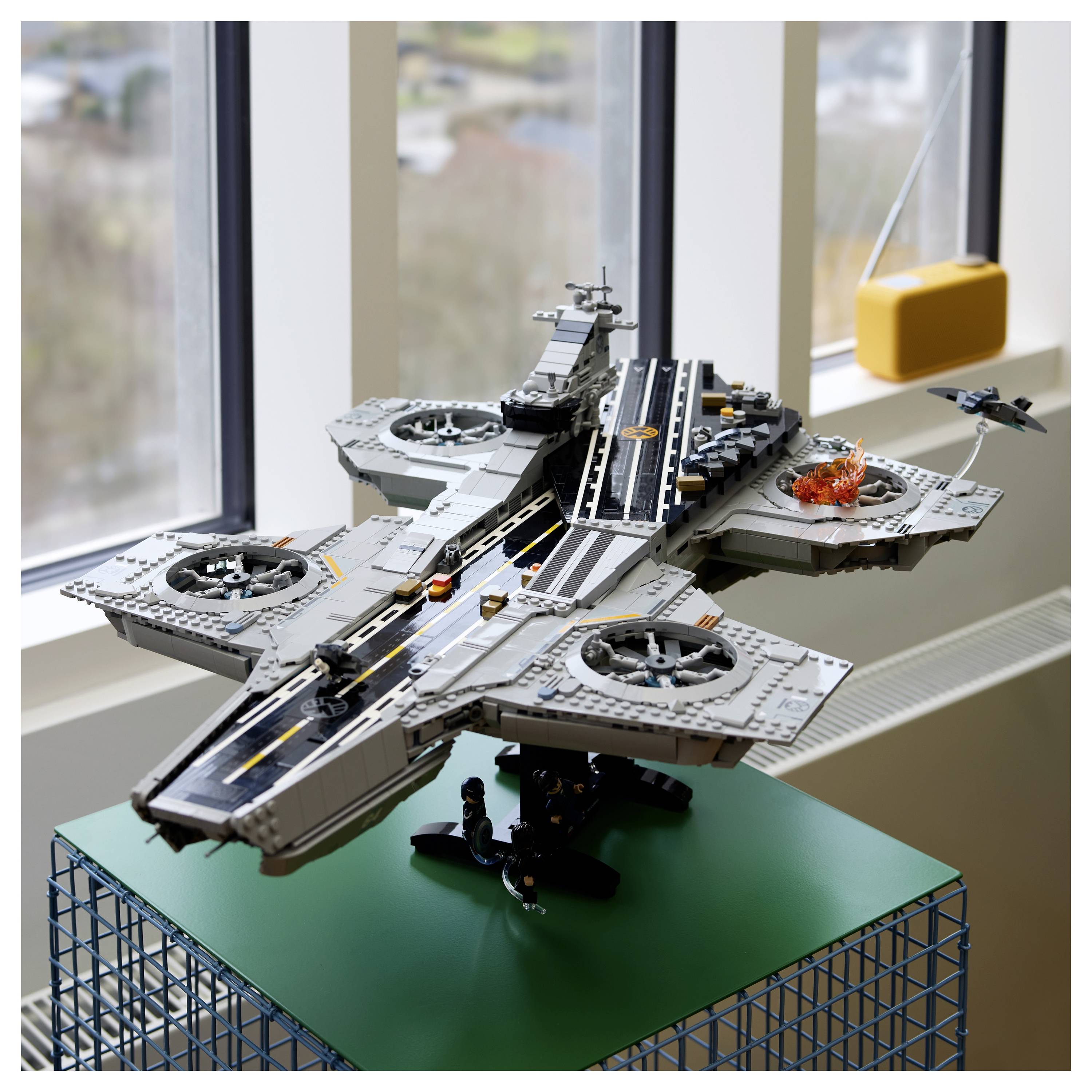 Ein detailliertes LEGO-Modell eines Flugzeugträgers auf einem grünen Ausstellungsständer, mit einer Landebahn, Hubschraubern, Düsenjägern und einer Explosionsszene.