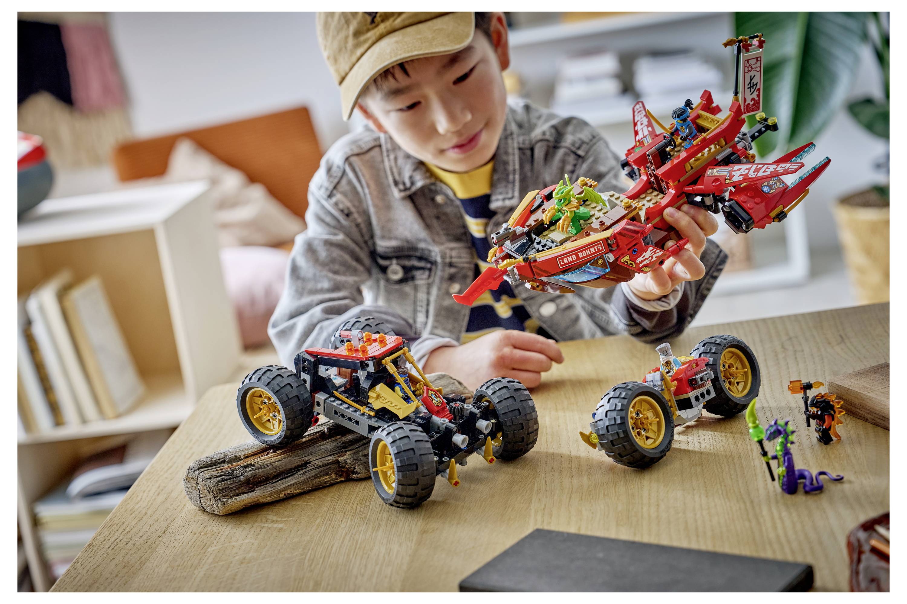 Kind untersucht ein rotes LEGO-ähnliches Flugzeugmodell an einem Tisch mit zwei kleinen Spielzeugfahrzeugen davor.