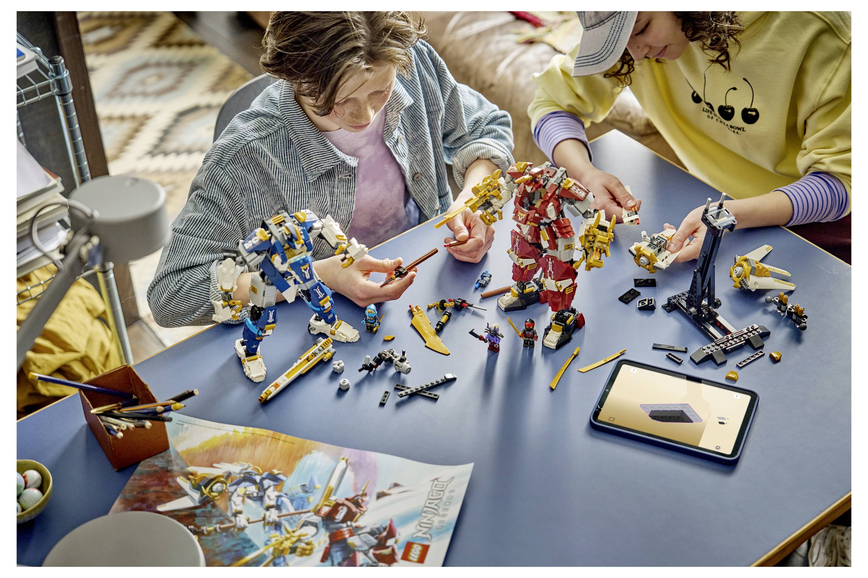Zwei Kinder bauen bunte Roboterfiguren aus einem LEGO-ähnlichen Bausatz an einem blauen Tisch, mit Anleitungen und einem Tablet in der Nähe.