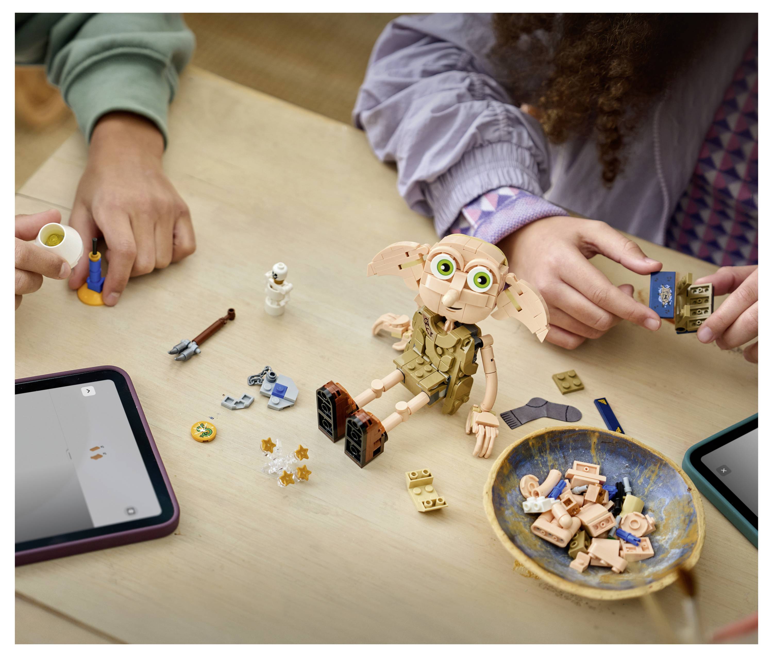 Kinder bauen ein LEGO-ähnliches Modell von Dobby auf einem Tisch zusammen, mit verstreuten Teilen, Tablets und einer Schüssel mit Bauteilen in der Nähe.