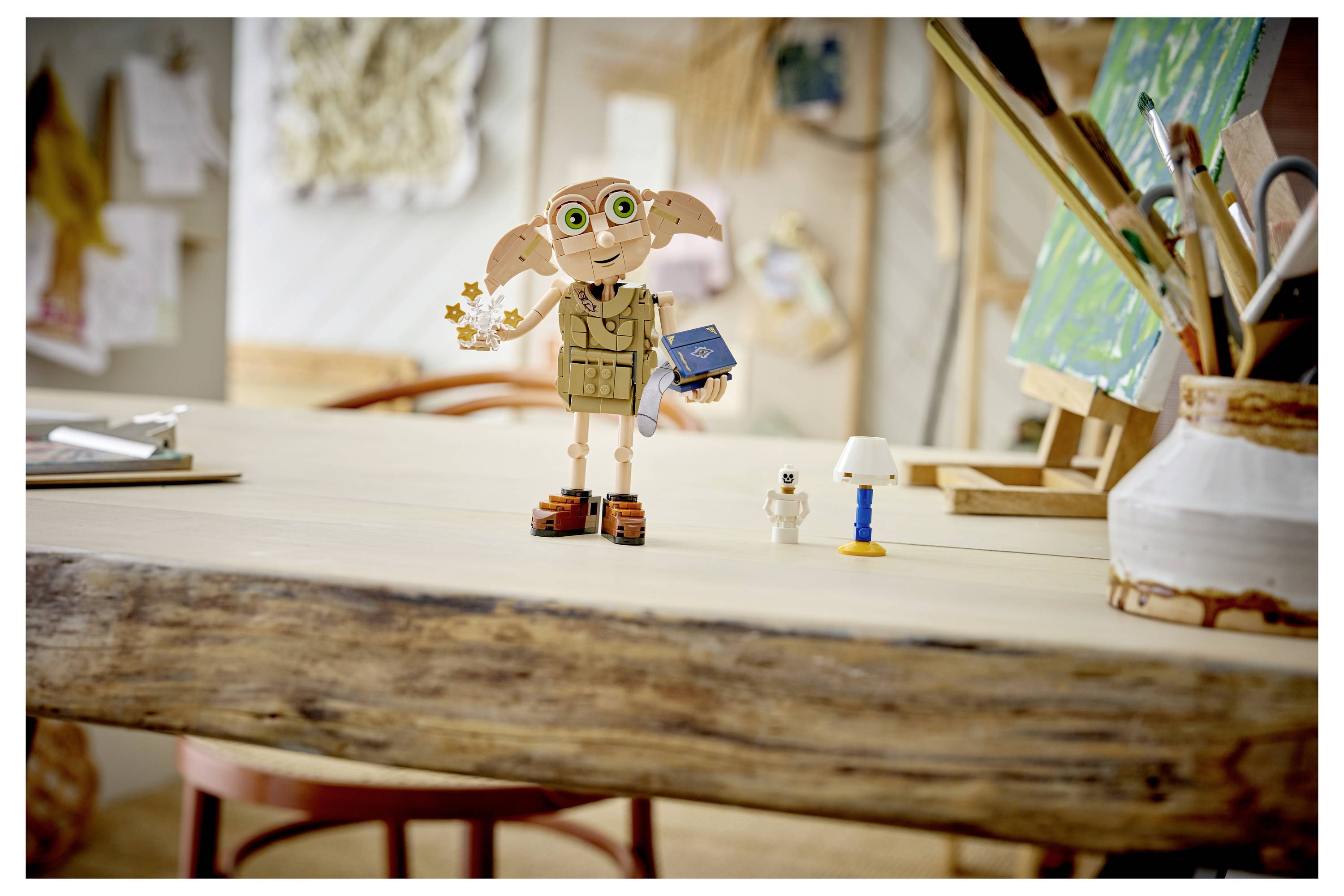 Eine kleine Dobby-Figur steht auf einem Holztisch in einem Kunststudio, hält ein blaues Buch und einen Zauberstab neben winzigen Schreibtischutensilien.