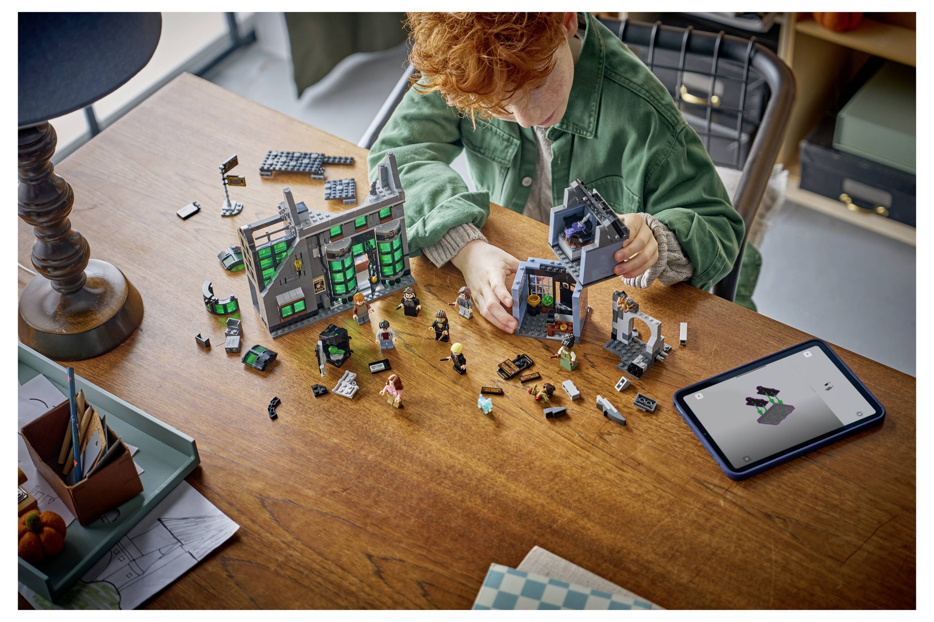 Ein Kind baut ein LEGO-Haus und einen Torbogen auf einem Holztisch auf, mit einem Tablet neben sich, das das Modell in den Anleitungen zeigt.