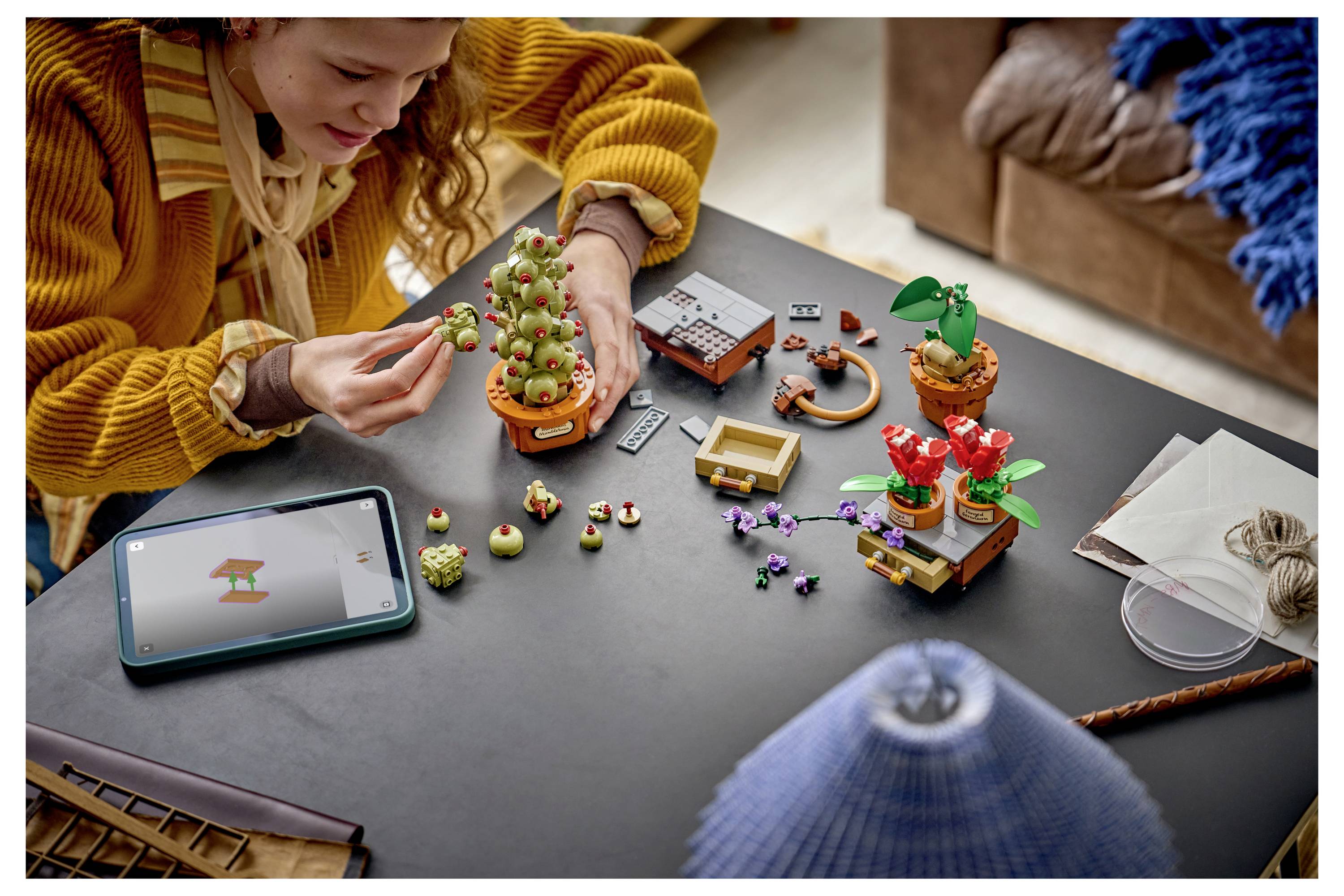 Eine Person, die an einem Tisch kleine LEGO-ähnliche Pflanzenmodelle zusammensetzt, mit einem Tablet in der Nähe, das Anweisungen anzeigt.