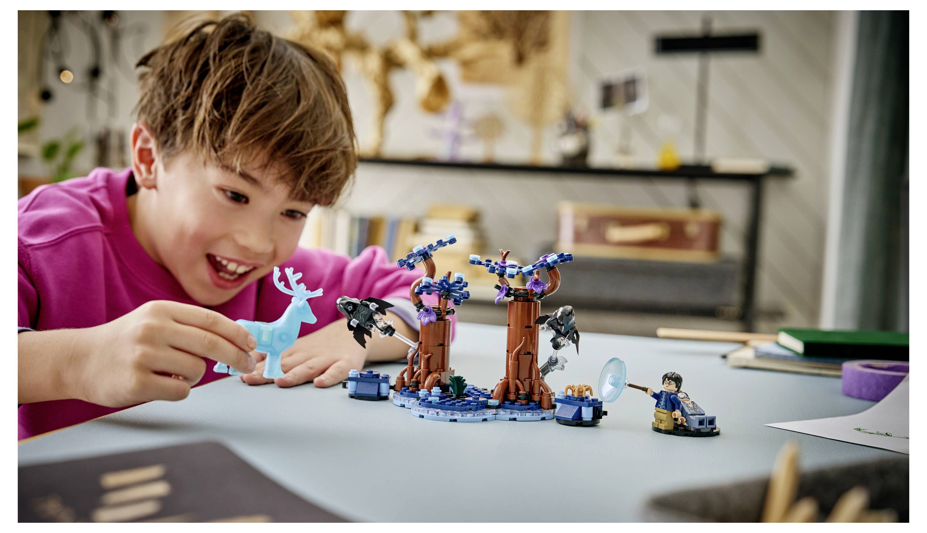 Junge in einem rosa Hemd, der lächelt, während er an einem Tisch mit einer LEGO-ähnlichen Fantasiebaumszene und blauen Figuren spielt.