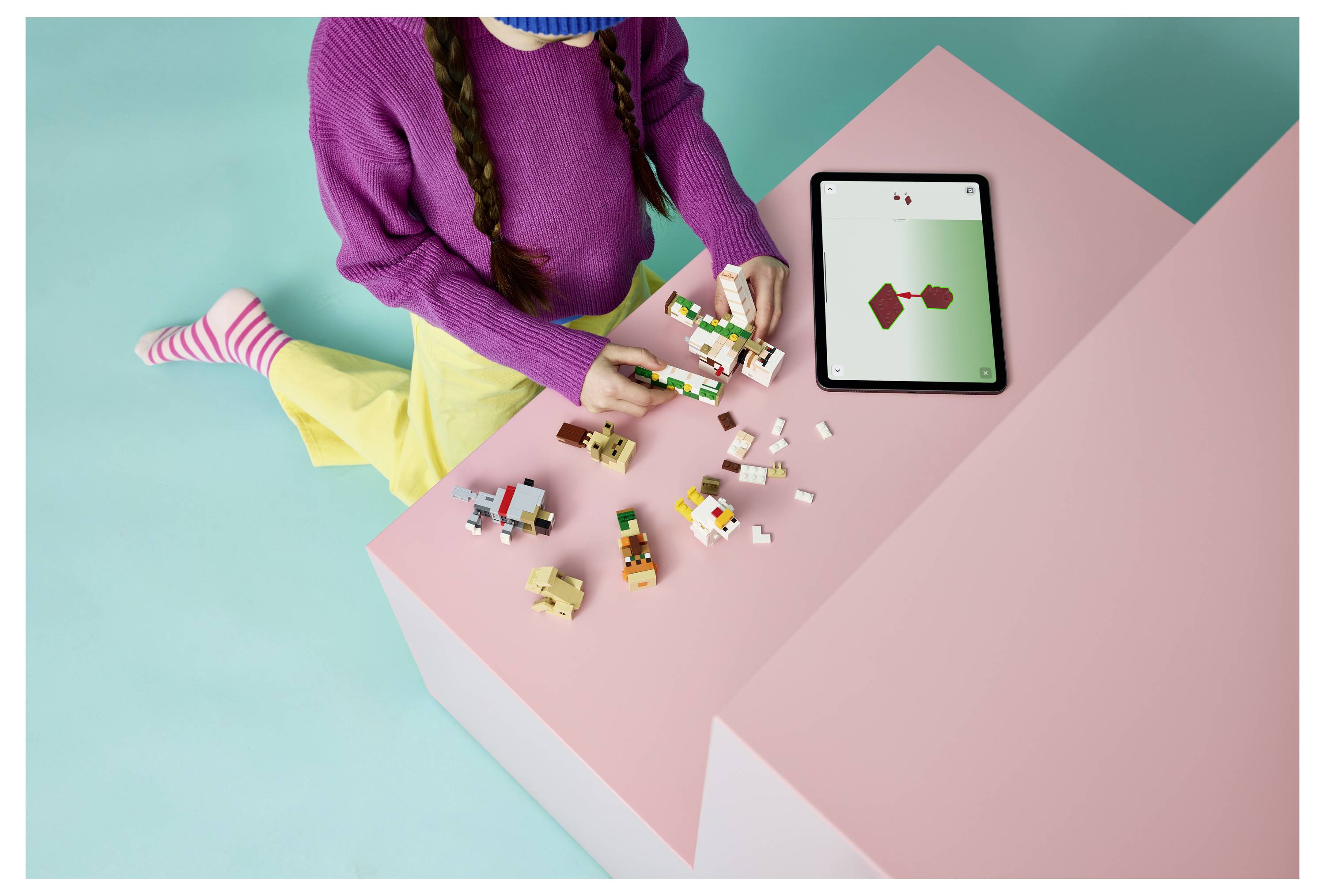 Ein Kind baut ein LEGO-Modell auf einem rosa Tisch neben einem Tablet, das ein einfaches Diagramm zeigt.