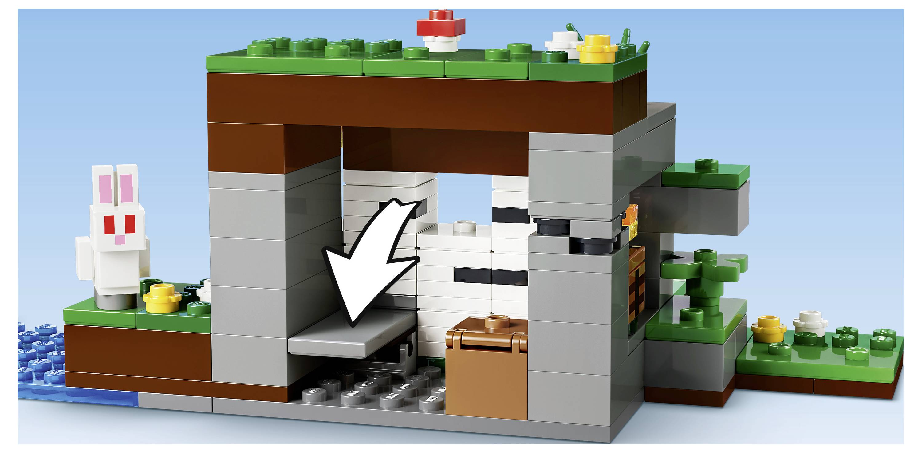 Ein LEGO-Höhleneingang mit einem weißen Pfeil, der auf eine versteckte graue Plattform im Inneren zeigt, neben einer braunen Truhe und einer weißen Kaninchenfigur außen.