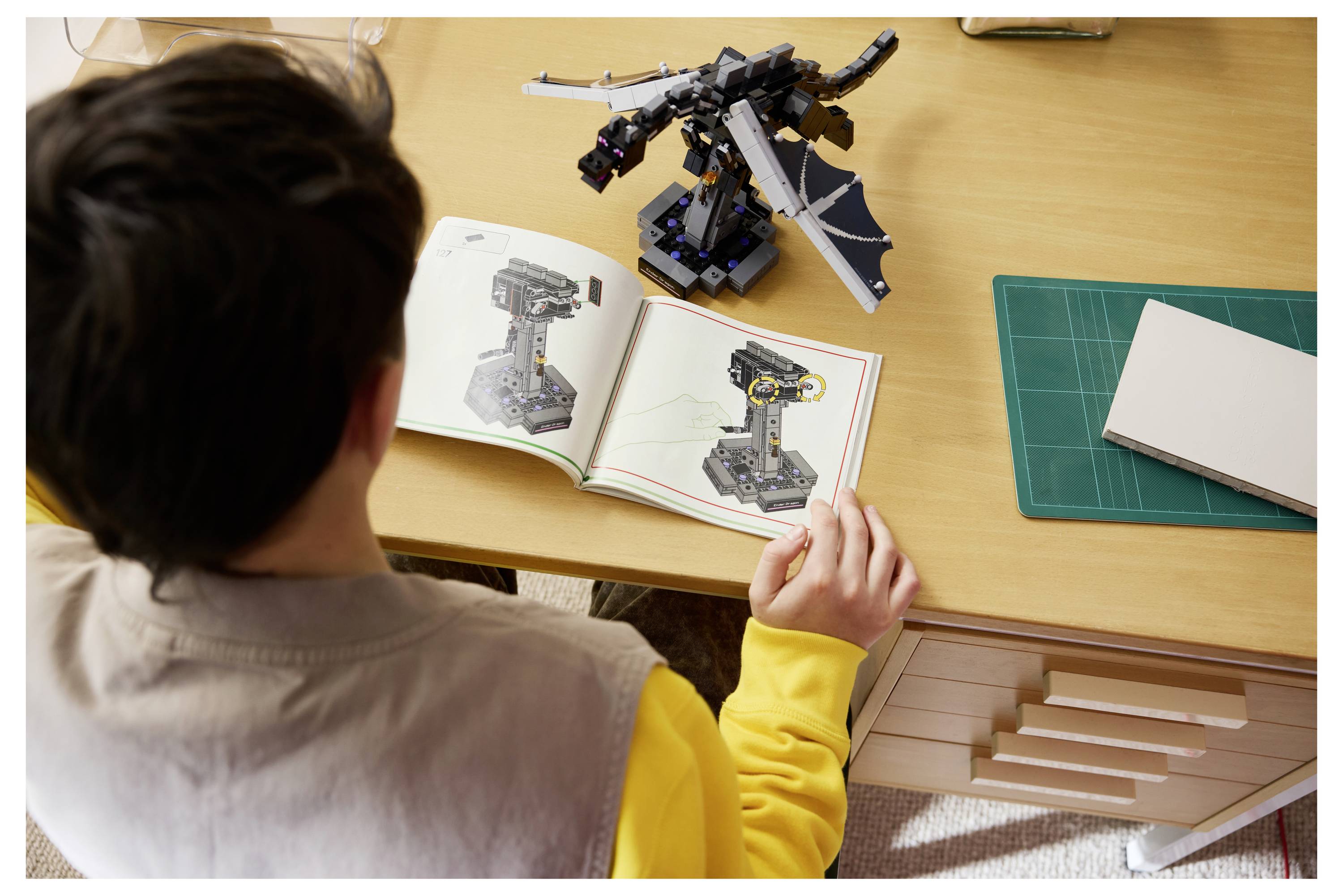 Person, die an einem Schreibtisch ein LEGO-ähnliches Modell baut und die zusammengesetzte drachenähnliche Figur mit Anweisungen in einem aufgeschlagenen Handbuch vergleicht.