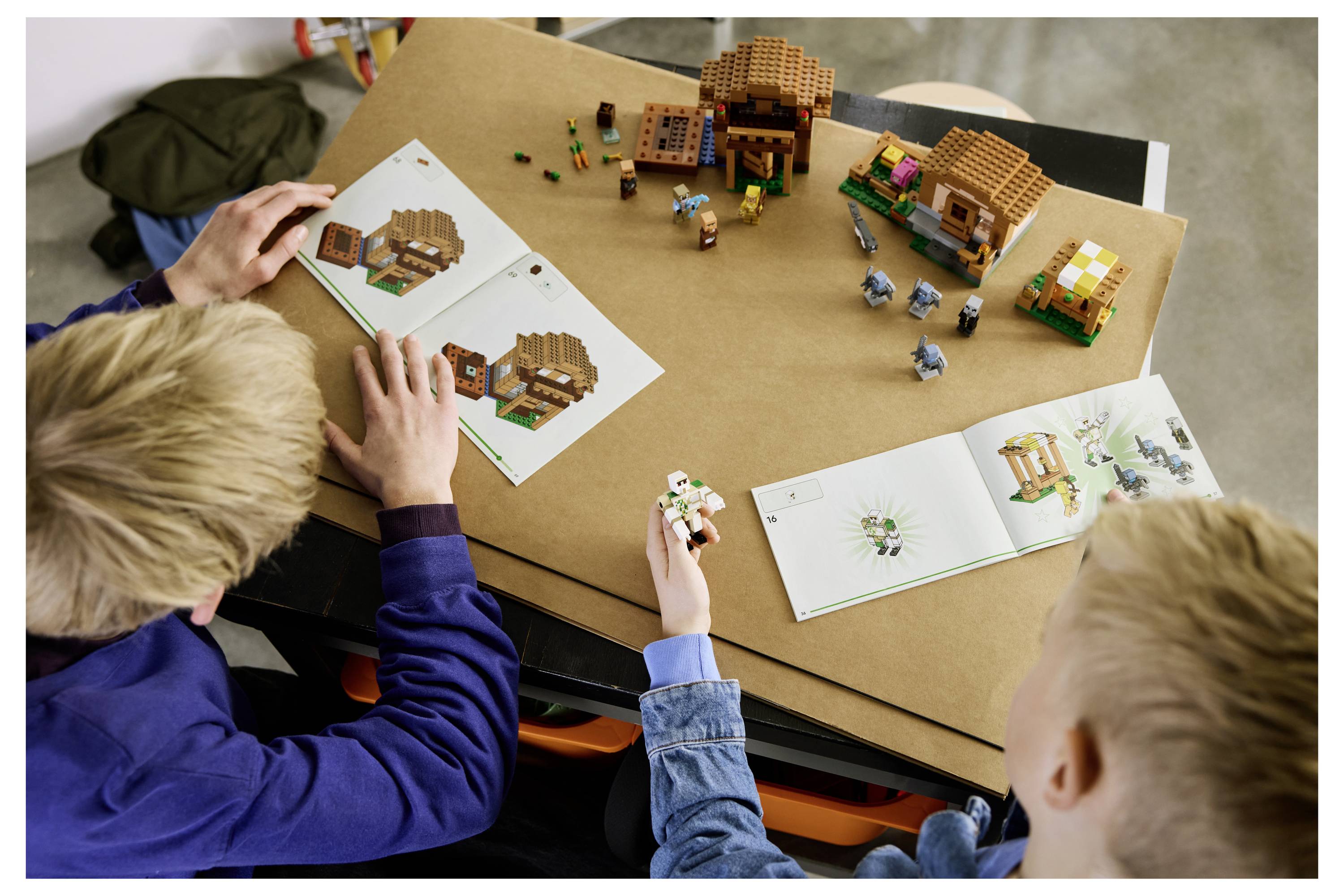 Zwei Kinder bauen ein LEGO-Dorf nach Anleituungsheften auf einem Tisch auf, mit kleinen Häusern und Figuren, die auf einer Pappunterlage angeordnet sind.