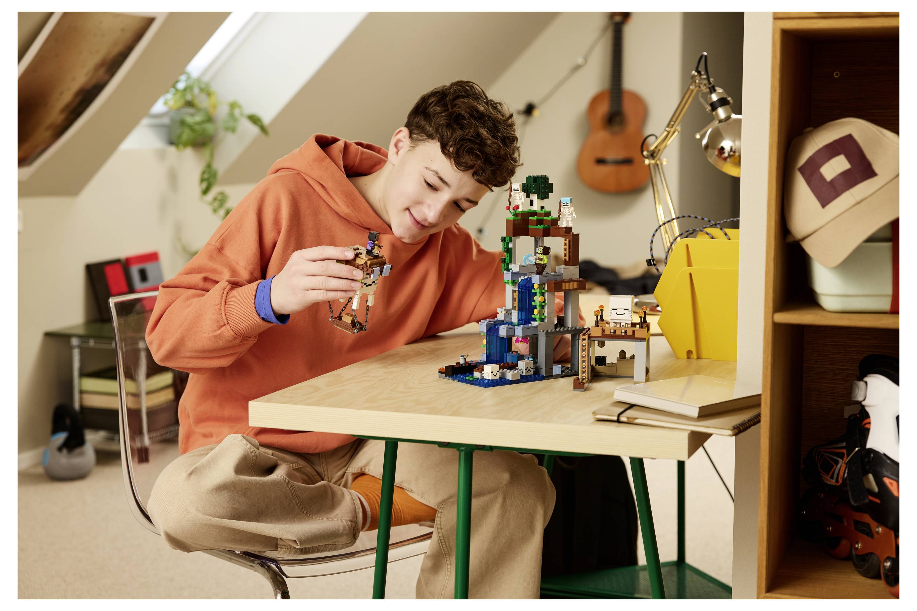 Teenager in orangem Hoodie, der an einem Schreibtisch in einem gemütlichen Zimmer ein LEGO-Modell baut und sich konzentriert auf eine kleine Figur neben einer größeren Ziegelmauer konzentriert.