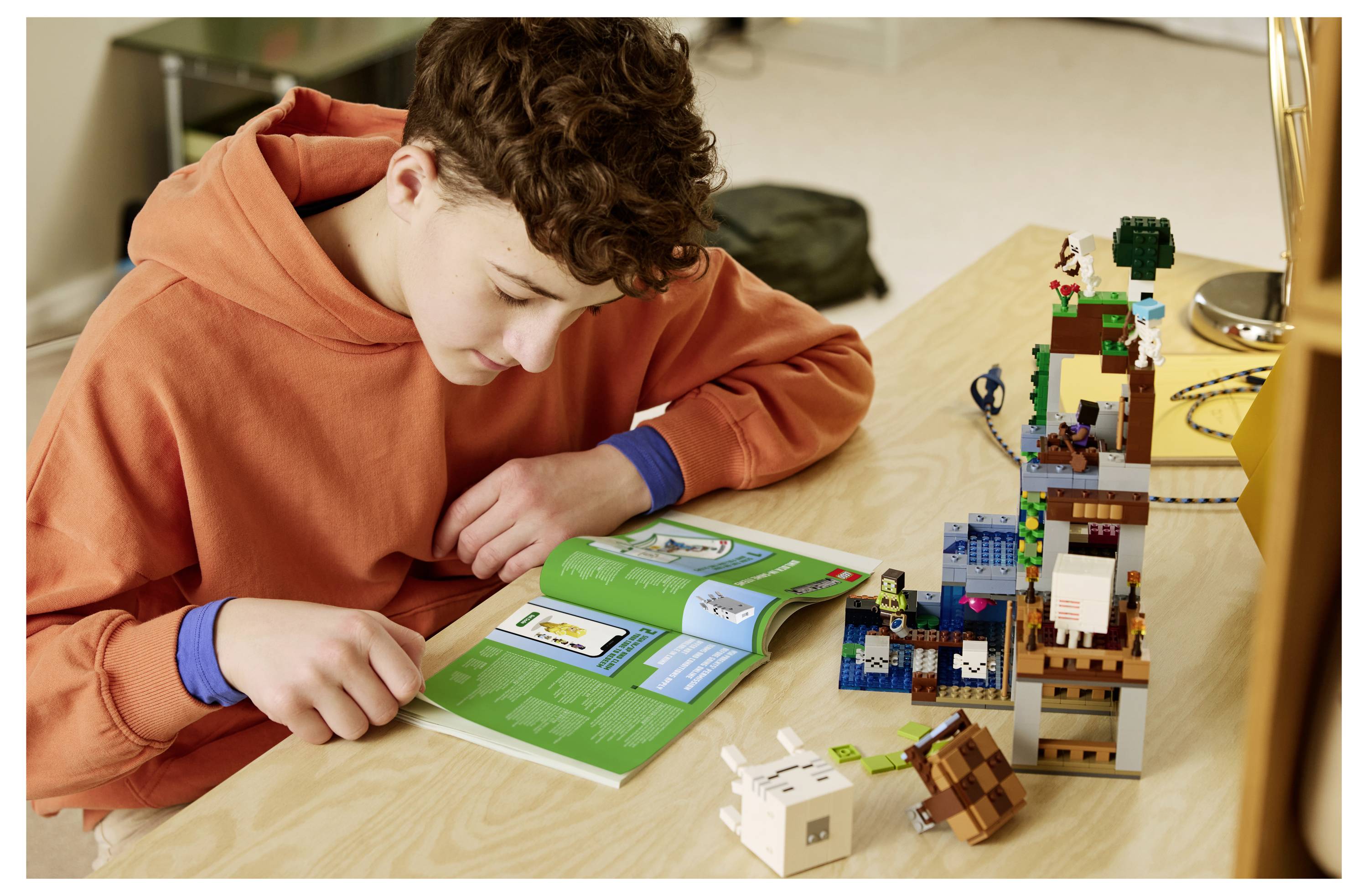 Junge in einem orangefarbenen Hoodie studiert ein offenes Anleitungsheft, während er einen LEGO-ähnlichen Turm auf einem Schreibtisch baut.