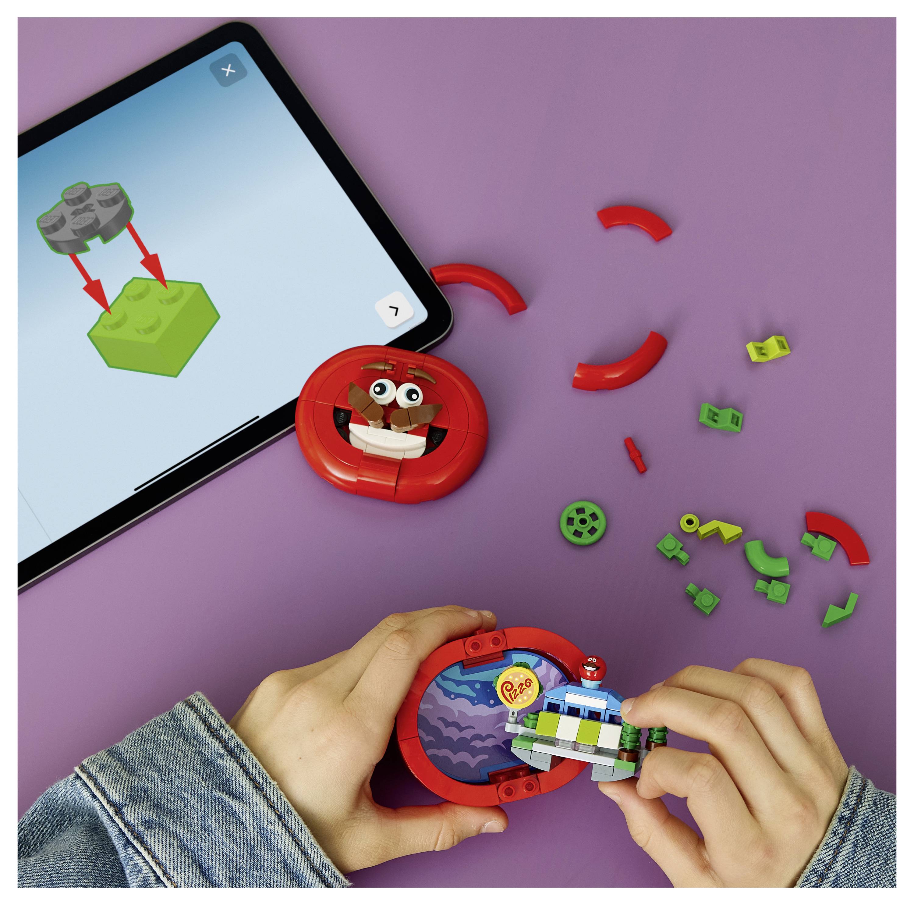Hände, die ein rotes kreisförmiges Spielzeug aus kleinen LEGO-ähnlichen Teilen zusammensetzen, während ein Tablet auf violettem Hintergrund Montageanleitungen anzeigt.