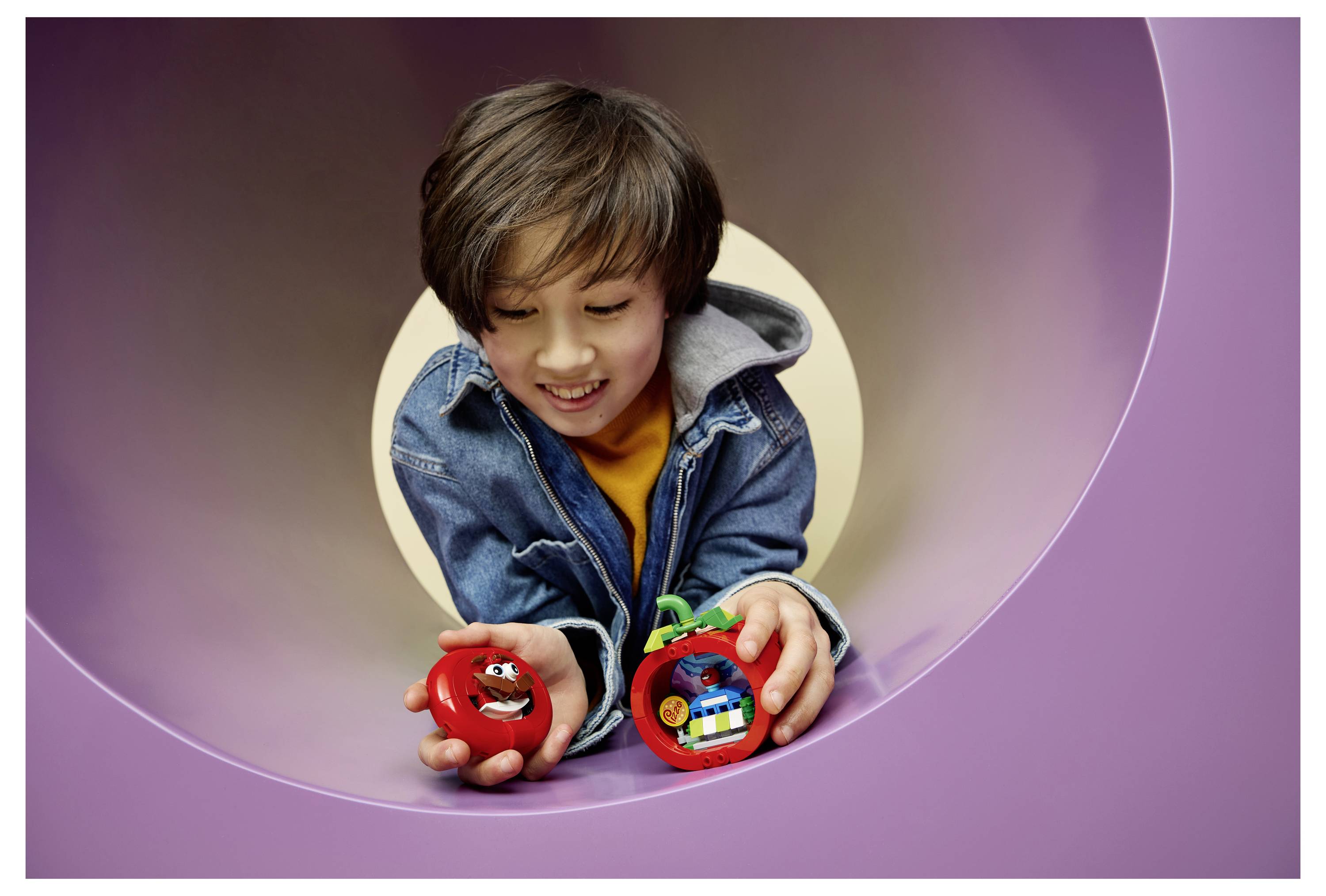 Ein lächelndes Kind in einer Jeansjacke hält zwei rote Spielzeugfahrzeuge in den Händen, während es durch eine violette Tunnelöffnung hindurchschaut.