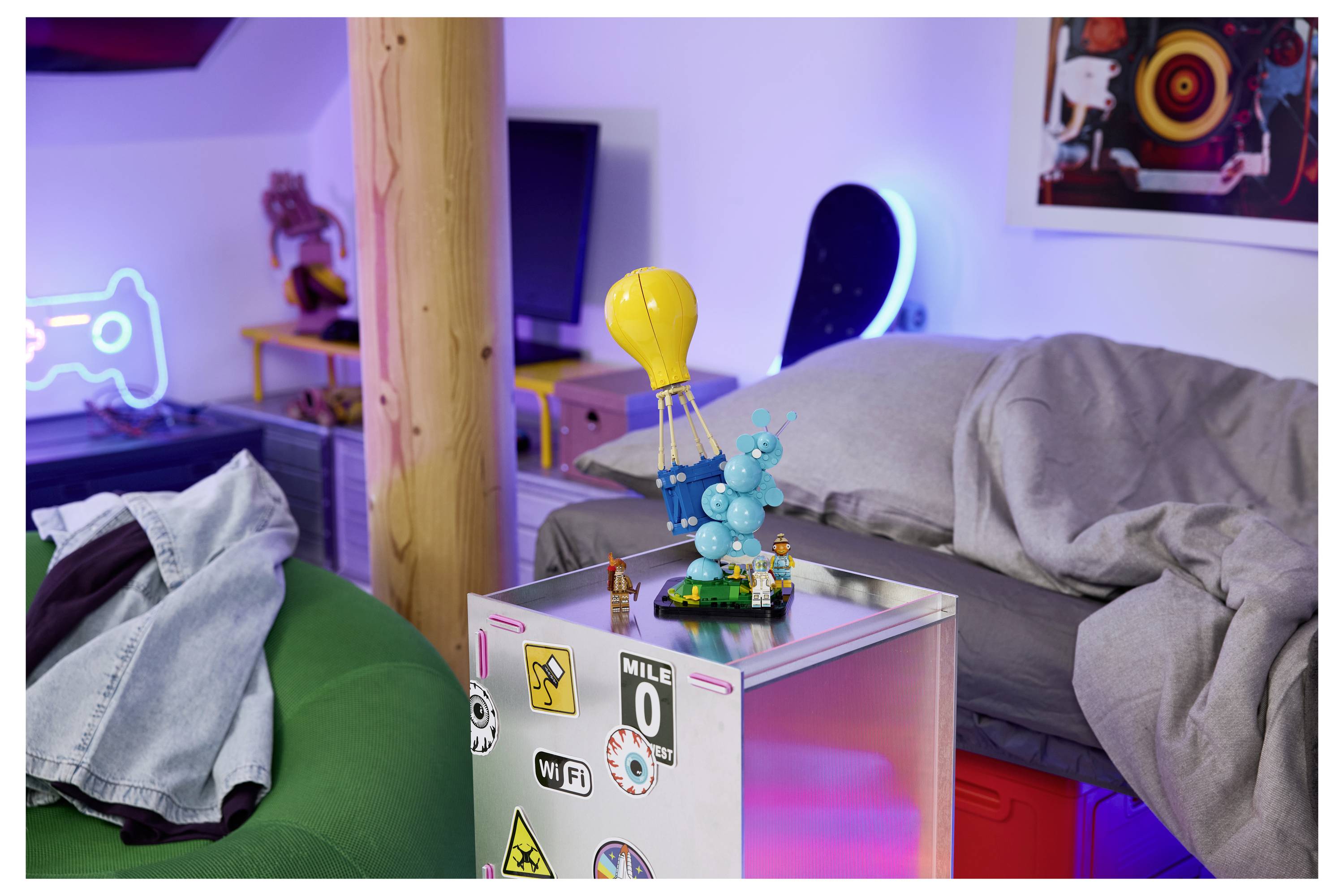 Ein gemütliches Schlafzimmer mit einem Bett, einem Sitzsack und einem mit Aufklebern bedeckten Mini-Kühlschrank, auf dem eine kleine dekorative Figur unter einer hängenden gelben Lampe steht.
