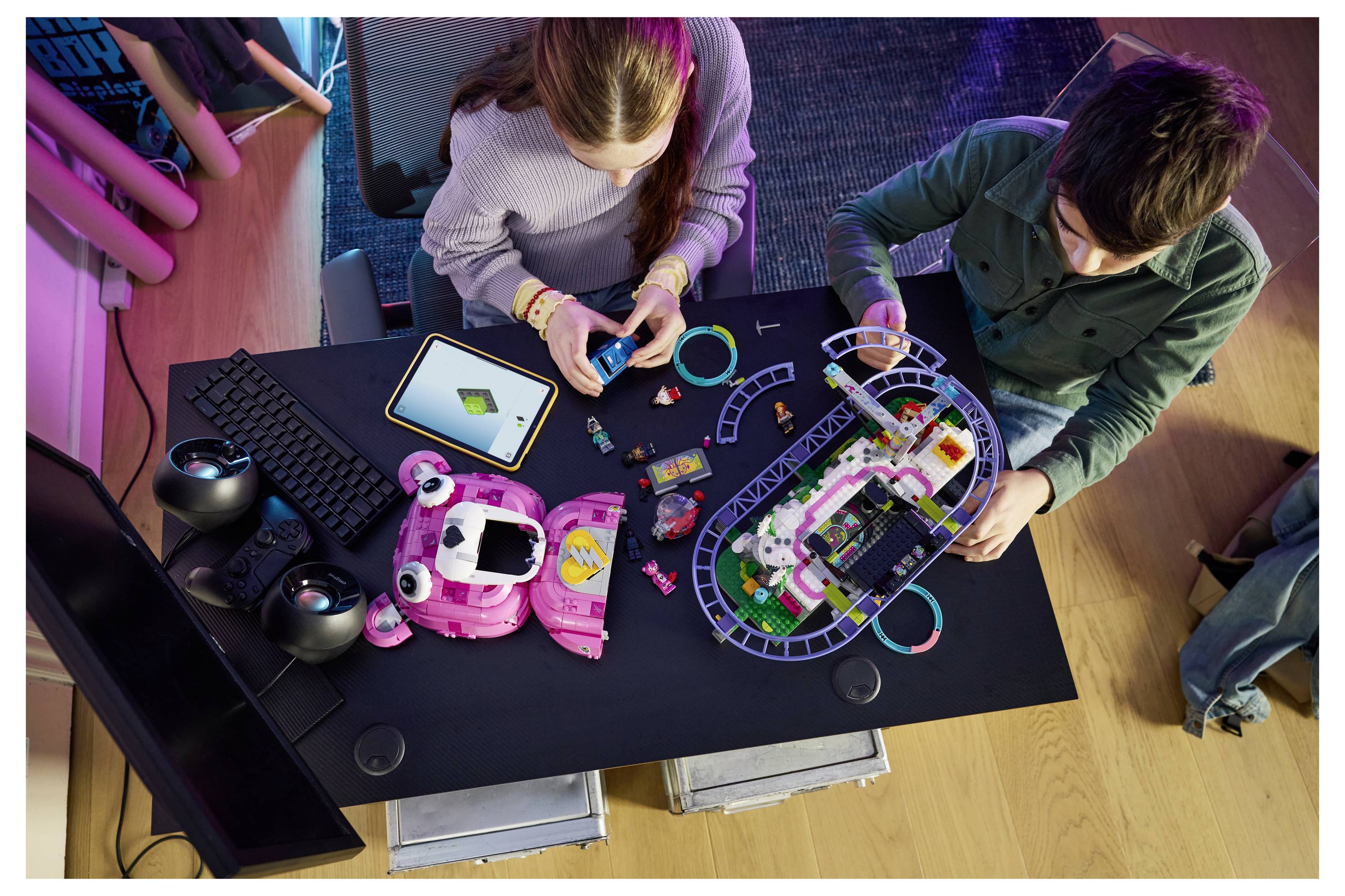 Zwei Kinder bauen ein farbenfrohen Spielzeugelektronik-Set auf einem schwarzen Tisch zusammen, mit Teilen, einem Tablet, einer Tastatur und Spielcontrollern in der Nähe.