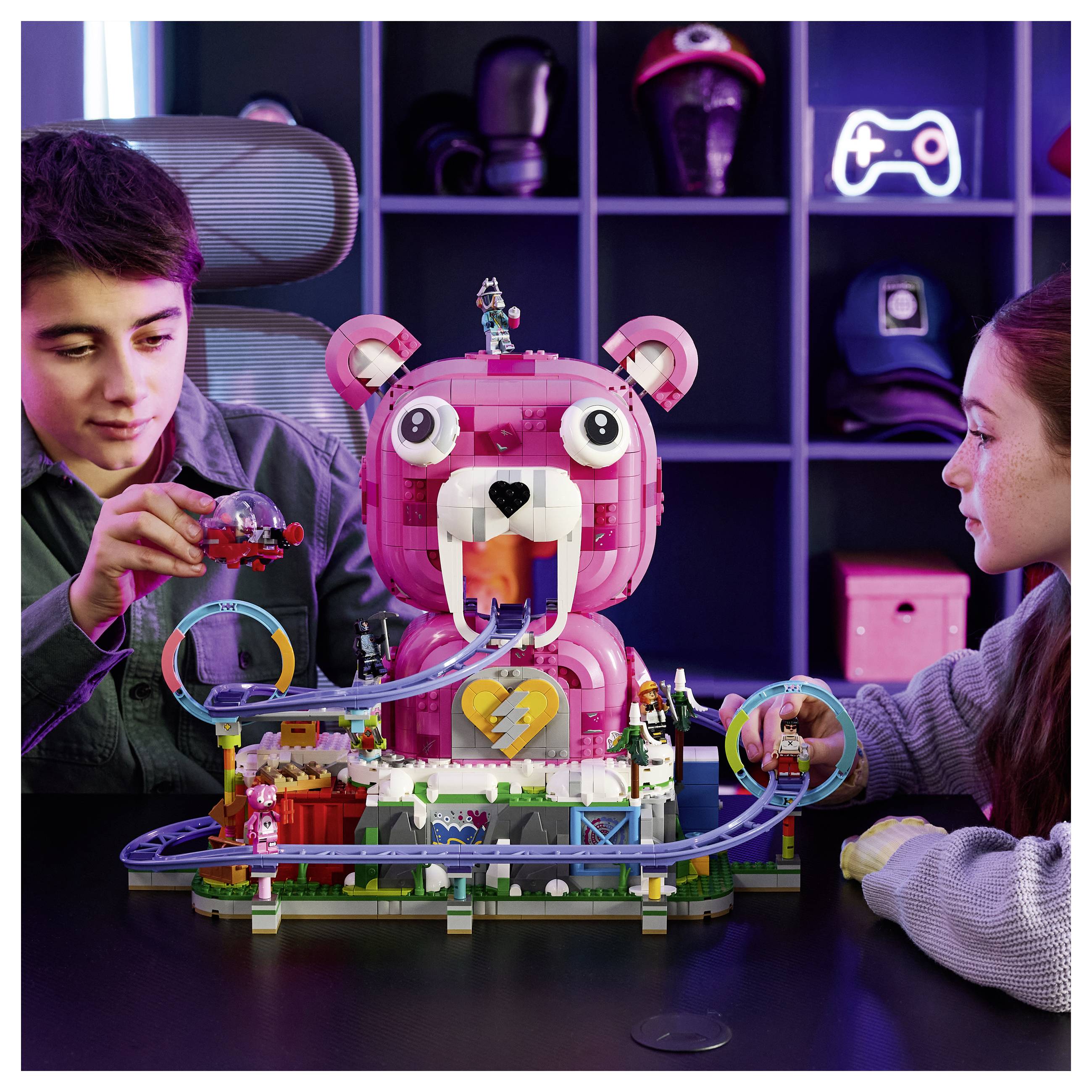 Zwei Kinder spielen mit einem rosafarbenen bärenthematischen LEGO-Achterbahn-Set an einem Tisch, mit einem großen Bärenmodell in der Mitte und einer Strecke, die sich um dieses herum schlängelt.