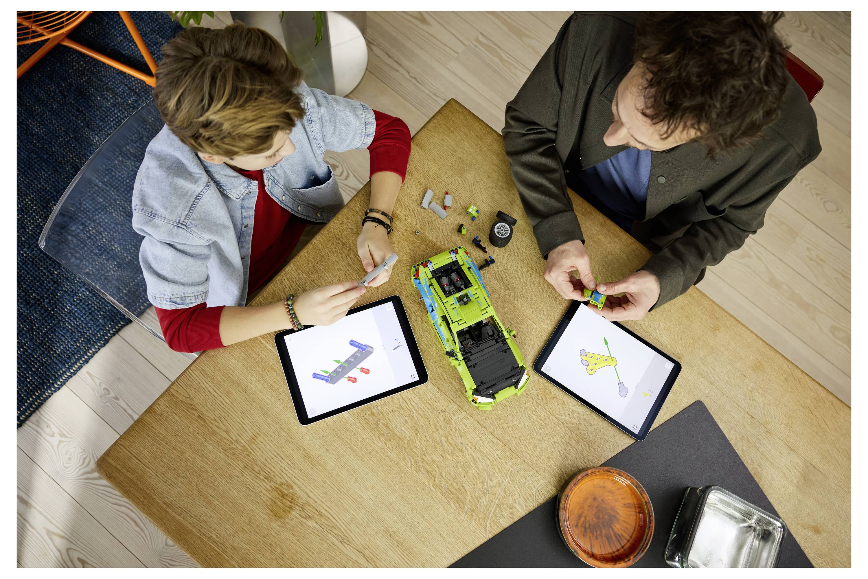 Zwei Personen montieren und inspizieren ein Spielzeugfahrzeug auf einem Tisch, wobei sie Tablets mit Teilediagrammen und nahegelegenes Werkzeug verwenden.