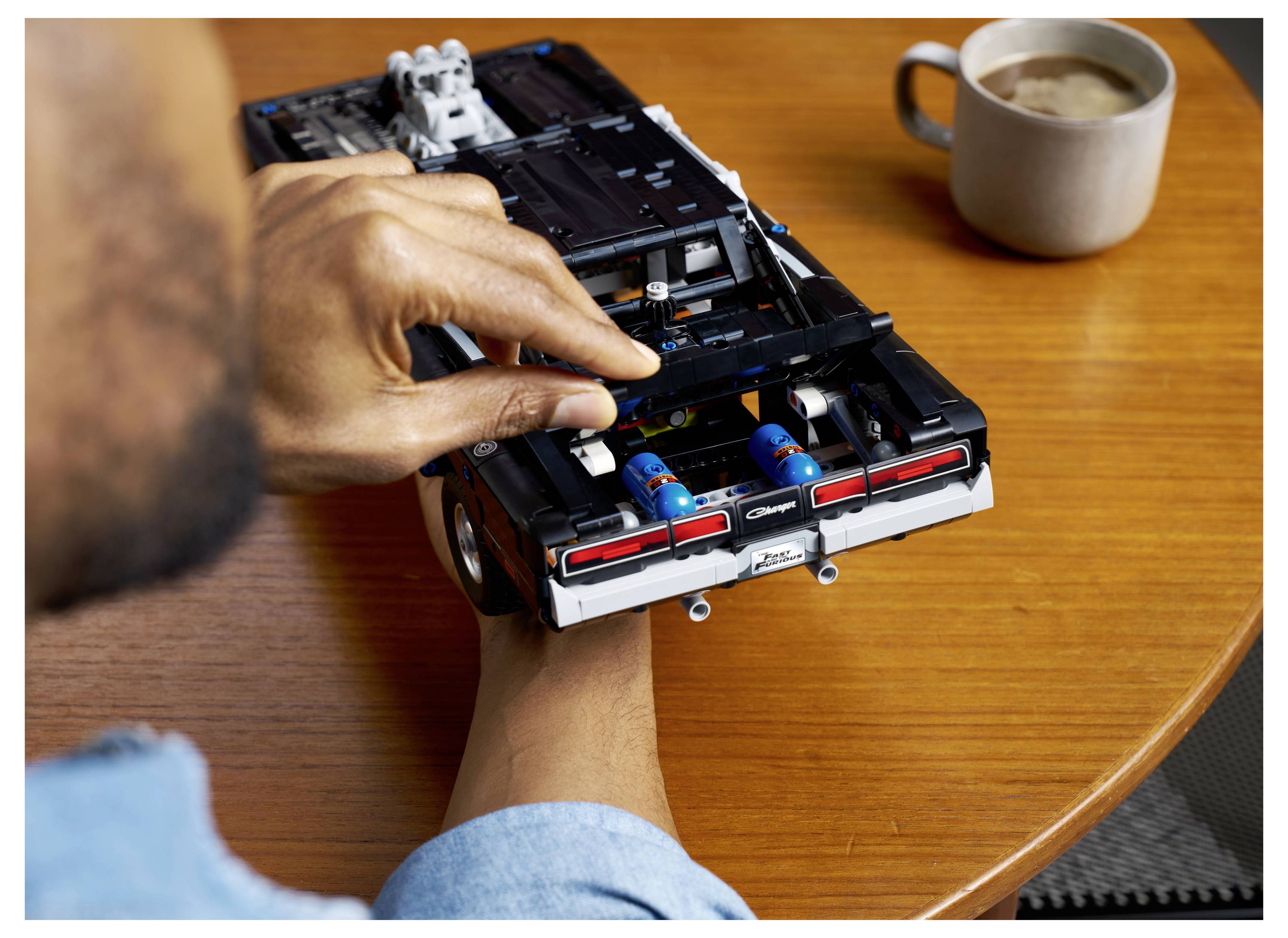 Eine Person setzt ein schwarzes LEGO-Automodell auf einem Holztisch neben einer Kaffeetasse zusammen.