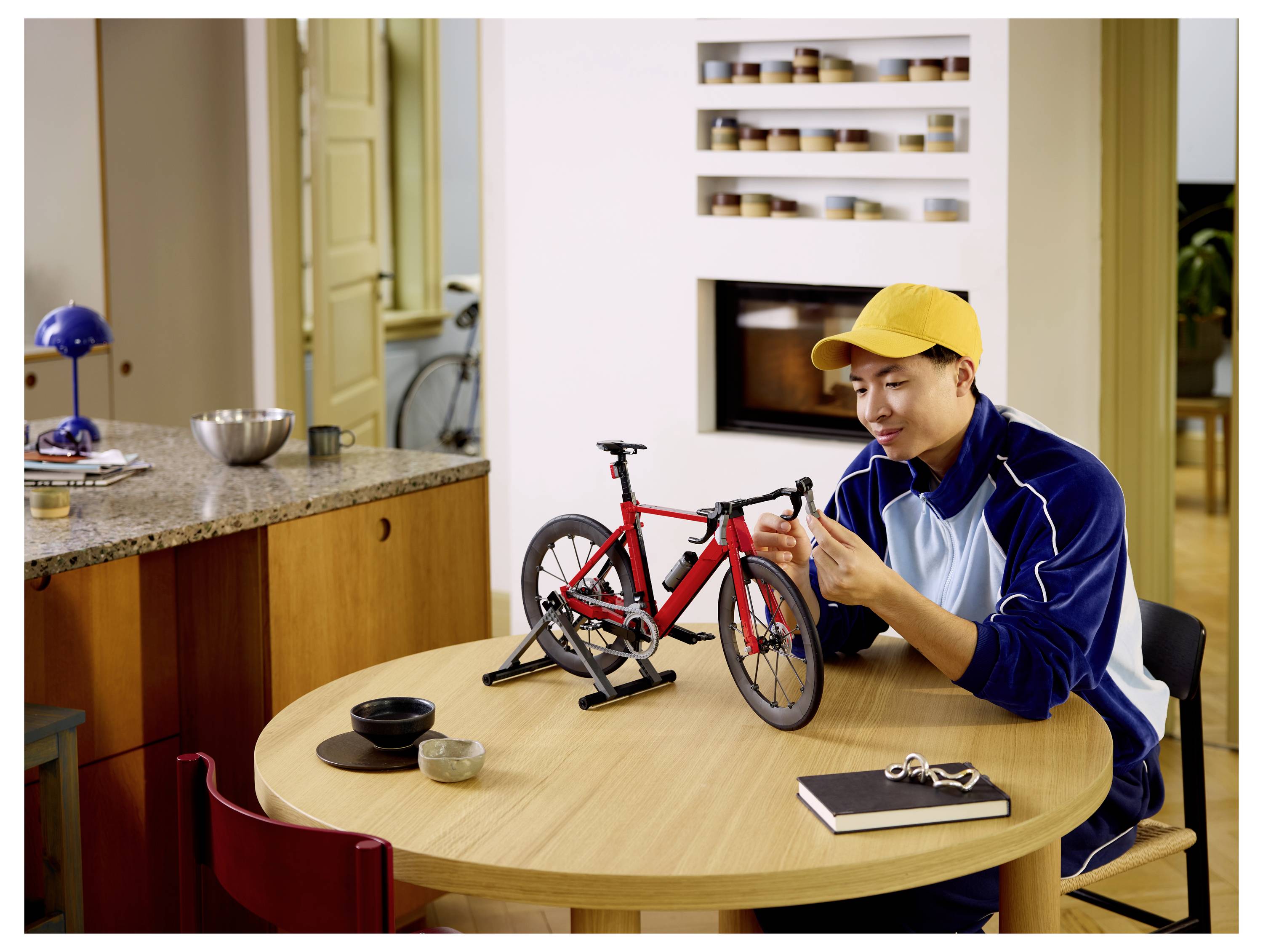 Ein Mann in einer gelben Kappe stellt ein kleines rotes Fahrradmodell auf einem Esstisch in einer hellen Küche ein.