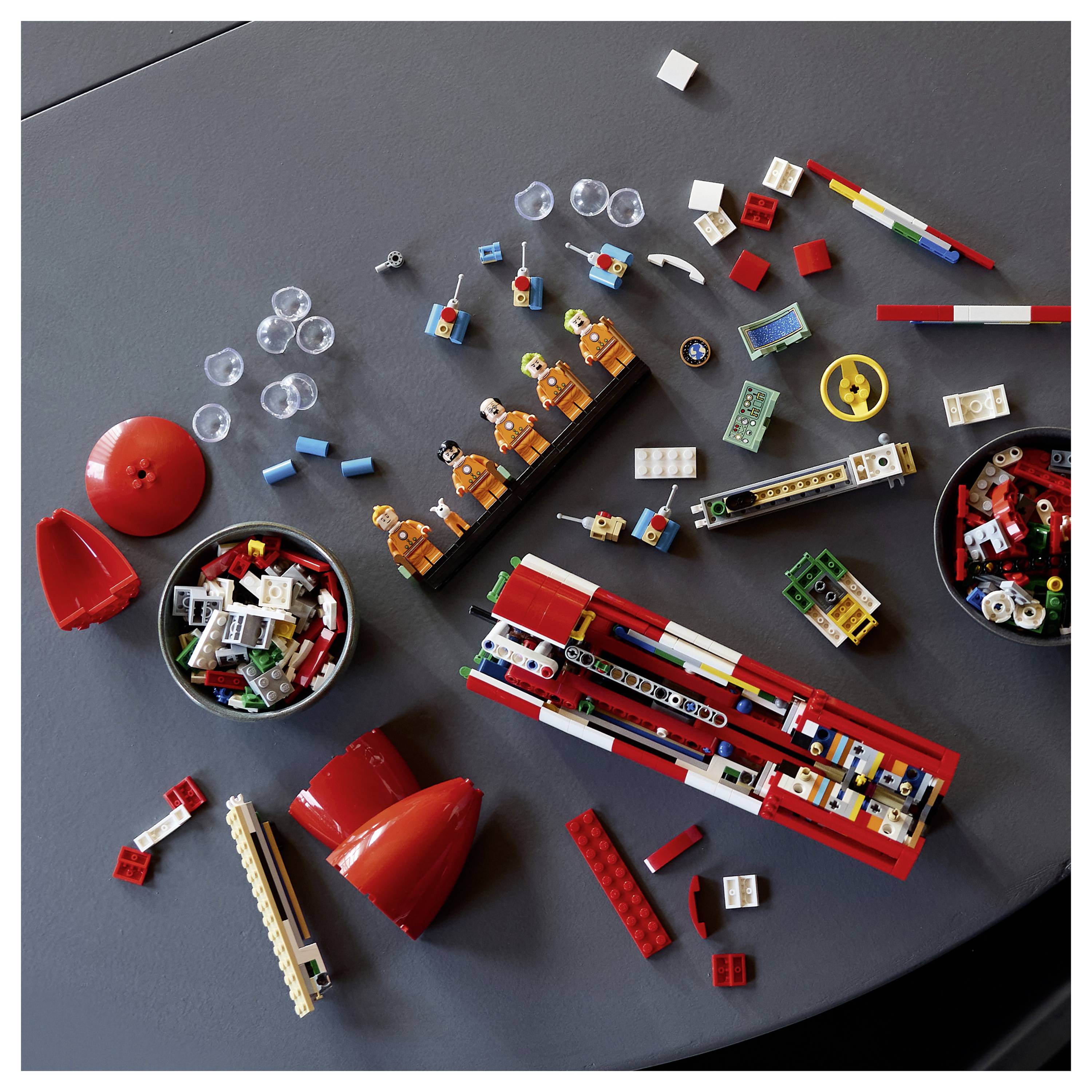 Verstreute LEGO-Teile und teilweise rote Modelle auf einem grauen Tisch, darunter Räder, Bausteine, Minifiguren und runde Behälter mit verschiedenen Teilen.