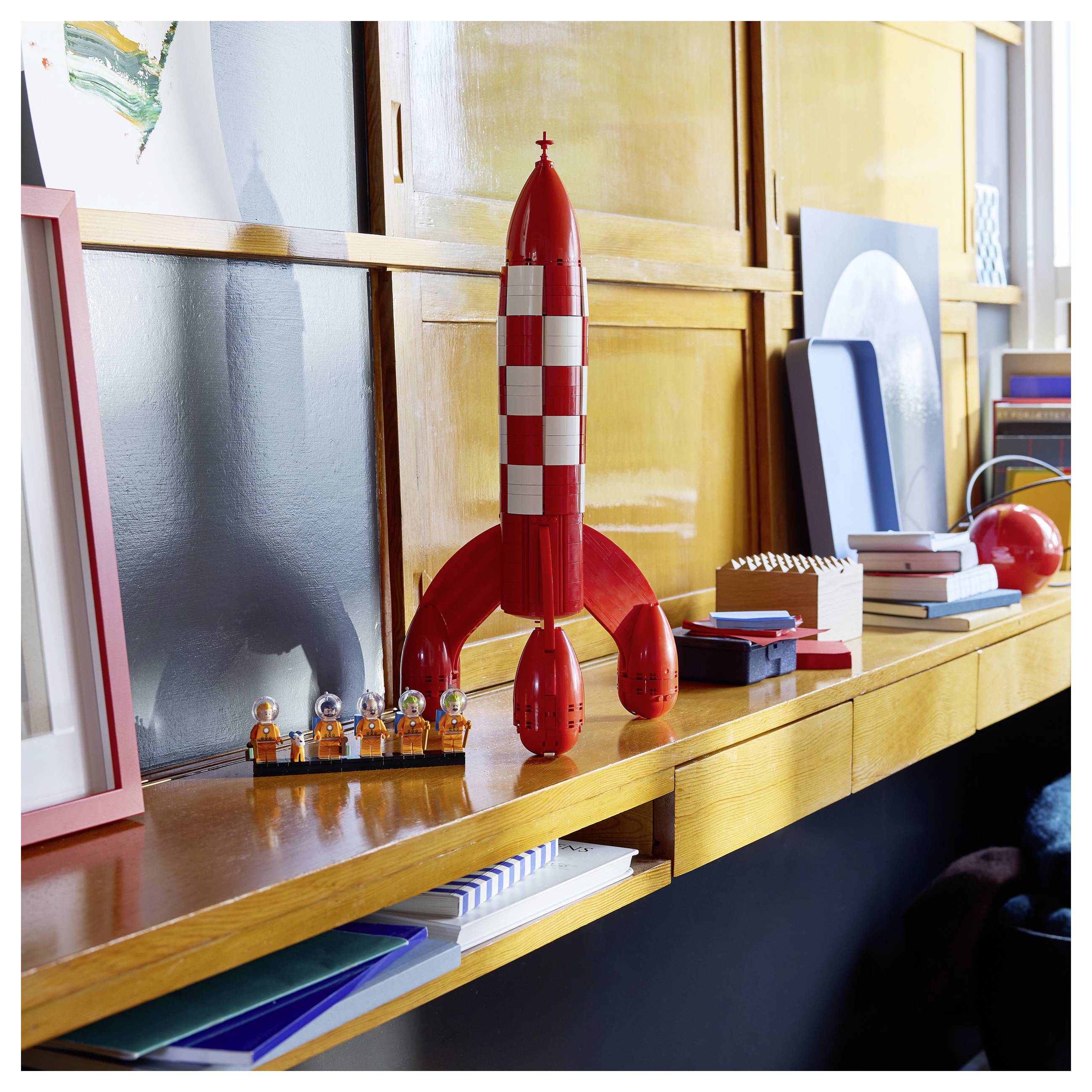 Rote Spielzeugrakete auf einem Holzregal, mit kleinen Astronautenfiguren und Büchern, in einem hellen Zimmer.