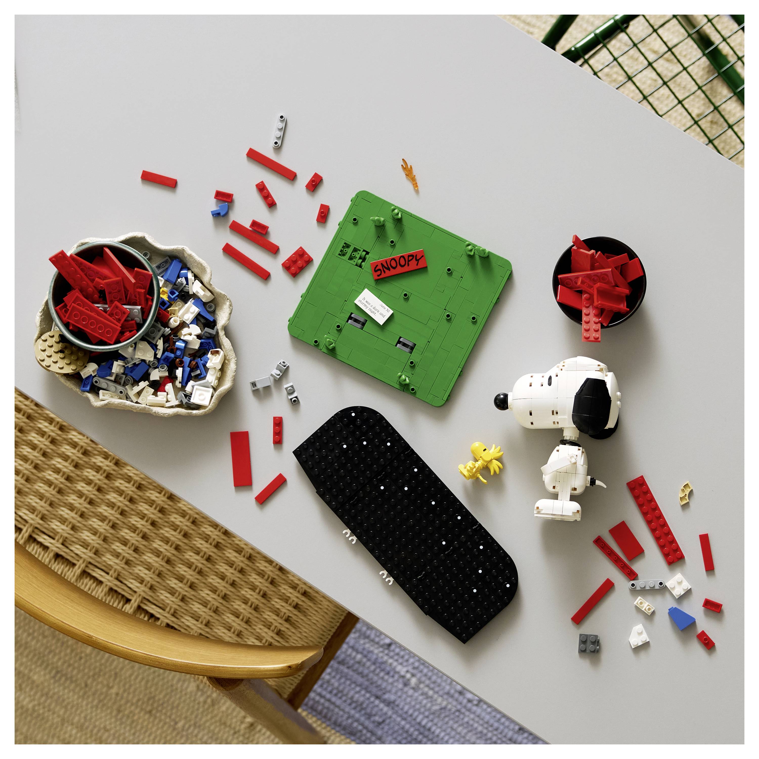 Draufsicht auf verstreute LEGO-Snoopy-Themen-Teile und Anleitung auf einem Tisch, mit roten, weißen, schwarzen und grünen Steinen und einer fertigen Snoopy-Figur