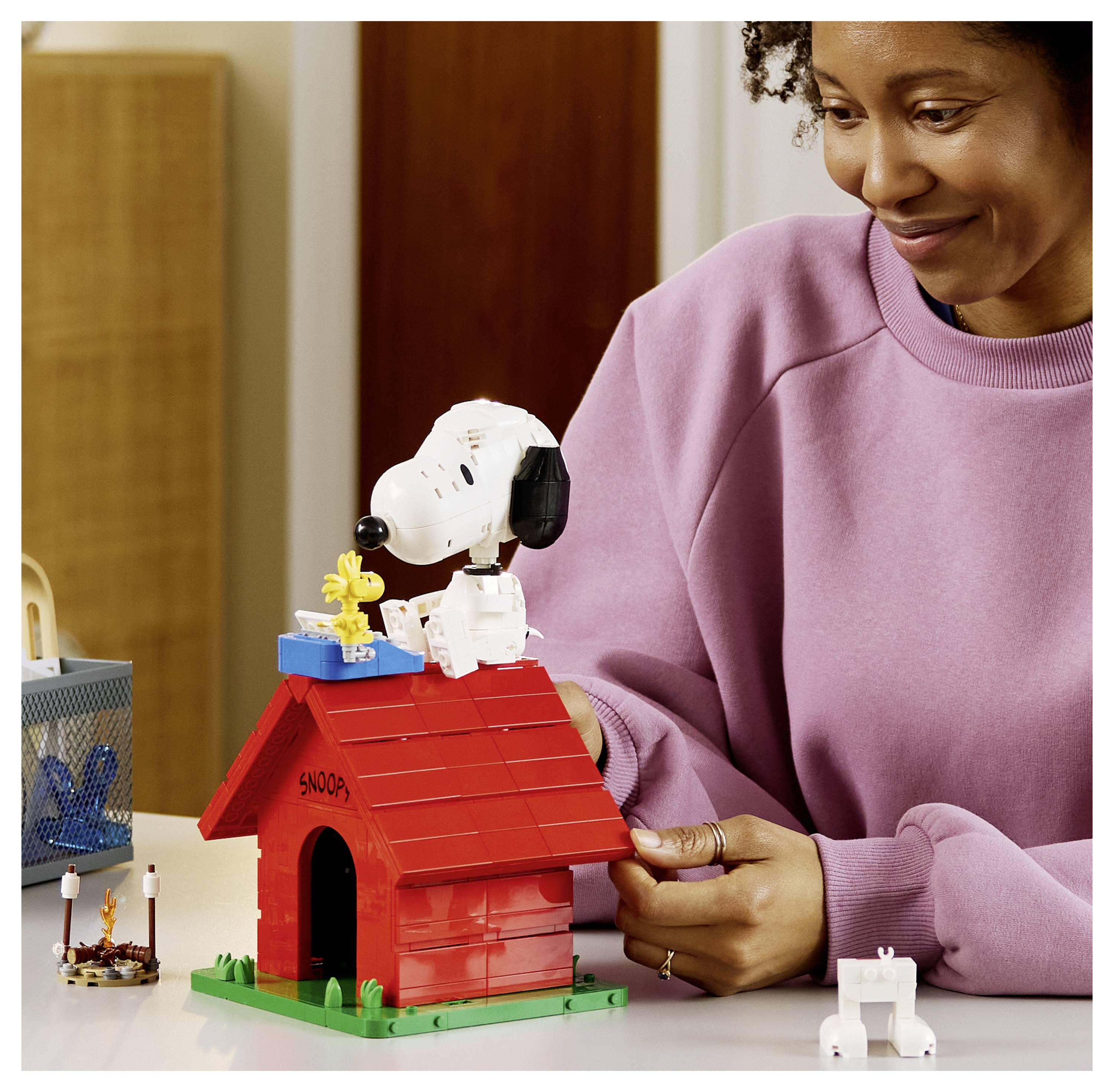 Eine Frau lächelt, während sie ein rotes Lego-Hundehaus mit einer Snoopy-Figur oben drauf baut, mit einer kleinen weißen Hundefigur in der Nähe.