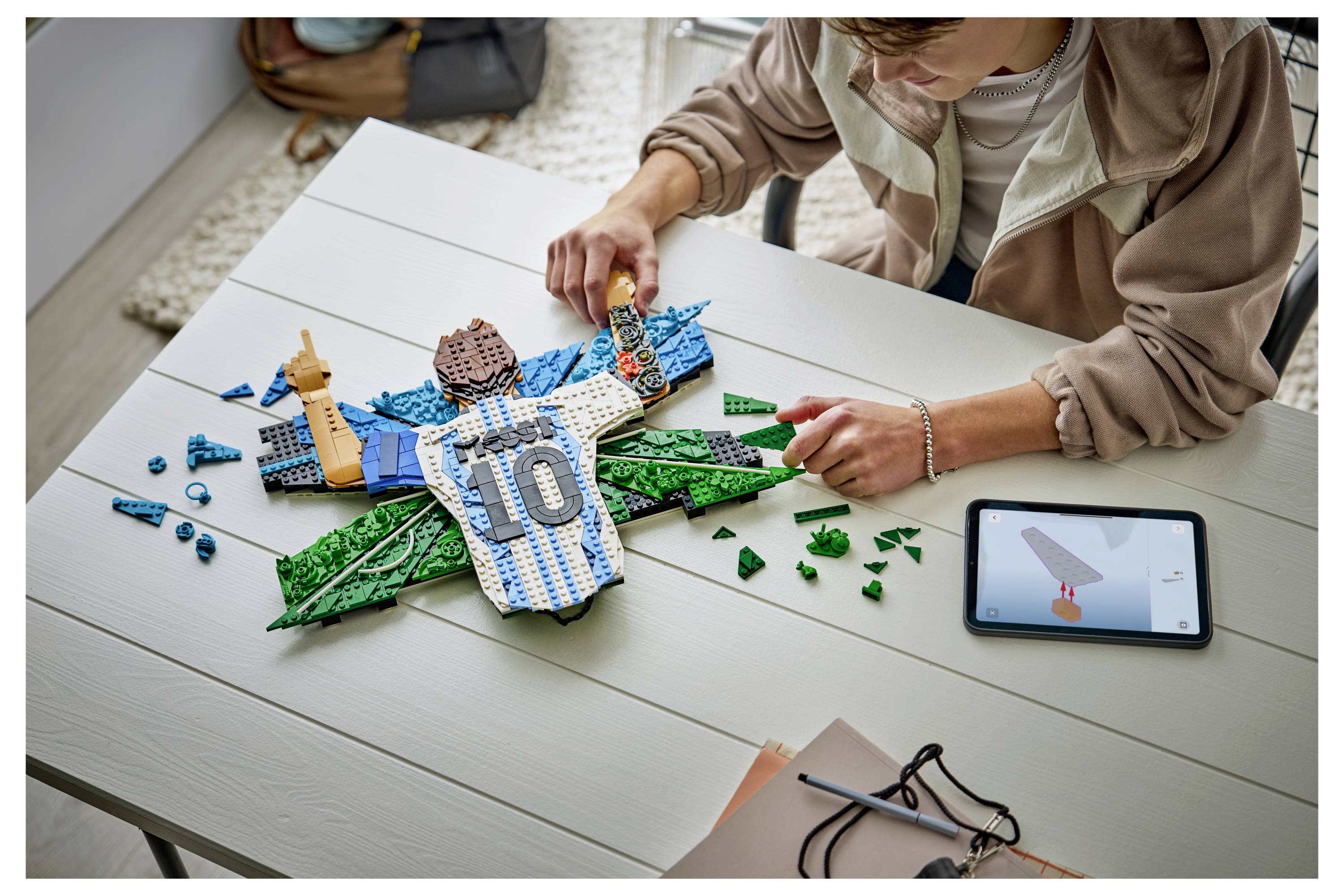 Person, die ein LEGO-Flugzeug auf einem Tisch zusammenbaut, wobei blaue und grüne Teile um einen Mittelteil im Trikot-Stil mit der Markierung 10 platziert werden, mit einem Tablet in der Nähe, das eine Rakete anzeigt.